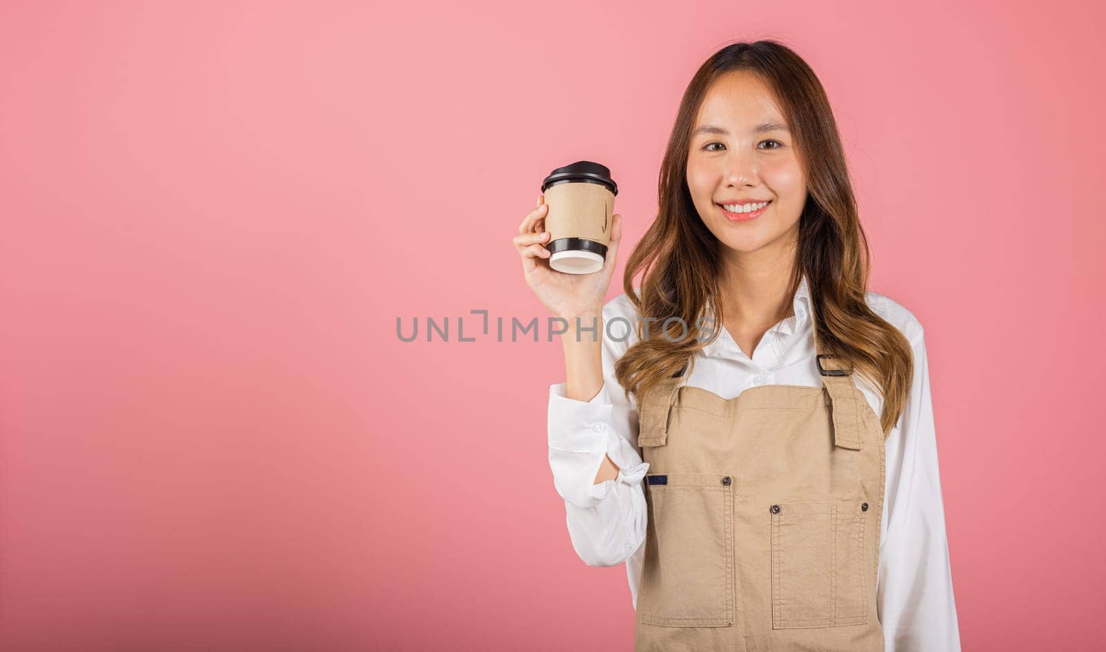 Portrait of Asian happy young woman barista bar tender coffee maker holding coffee tea hot cup studio shot isolated on pink background, female person smiling hold takeaways cup by Sorapop