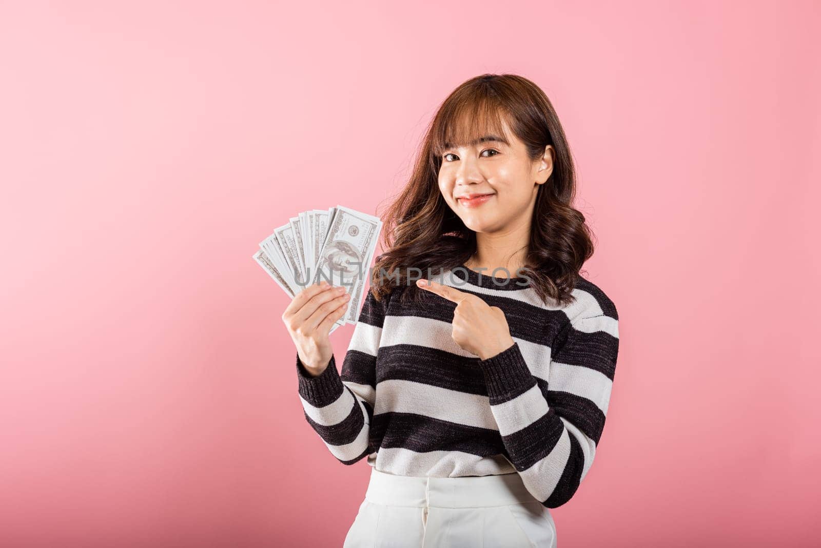 Smiling face portrait asian beautiful woman cheerful her holding and point the money fan banknotes 100 dollar bills on pink background, with copy space for text