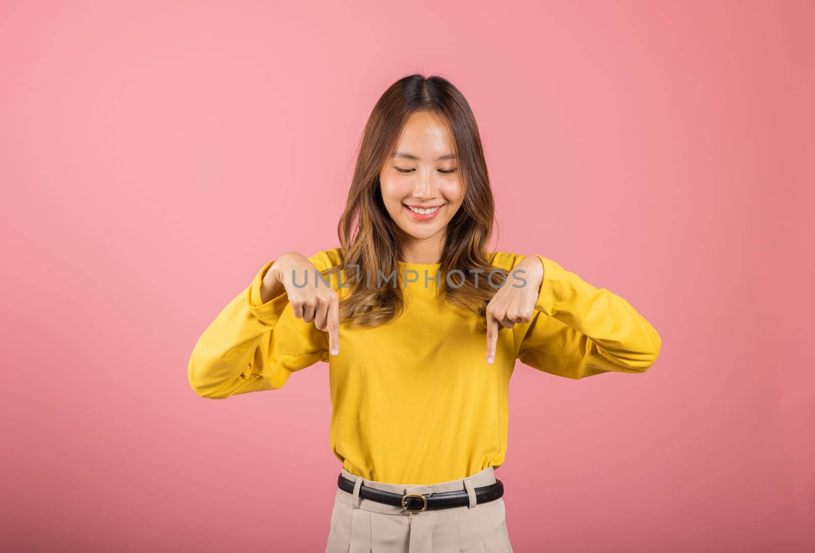 Asian happy portrait beautiful young woman smiling makes gesture two fingers pointing below down presenting and looking, studio shot isolated on pink background with copy space, point down