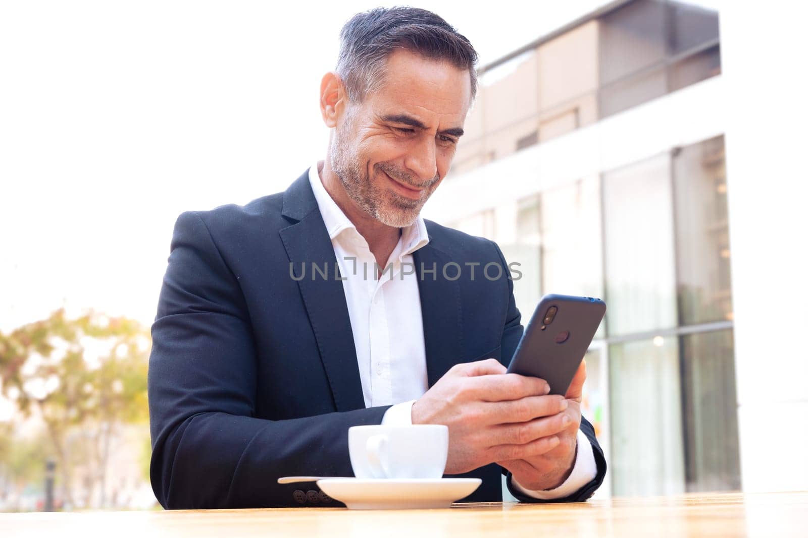 Satisfied with the results, the businessman sitting outside the office building, a mature boss holds a phone and drinks coffee, writes messages and reads news online, using an app.