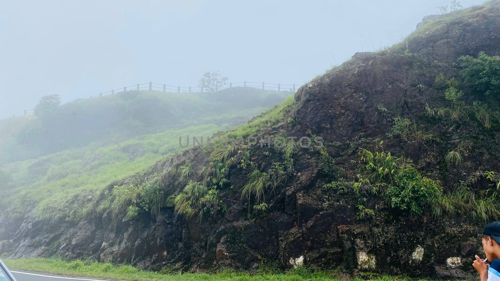 A scenic view of a misty mountainside with lush vegetation and a person standing near a road, taking in the beautiful scenery. The fog blankets the top of the hill, creating a sense of mystery and tranquility.