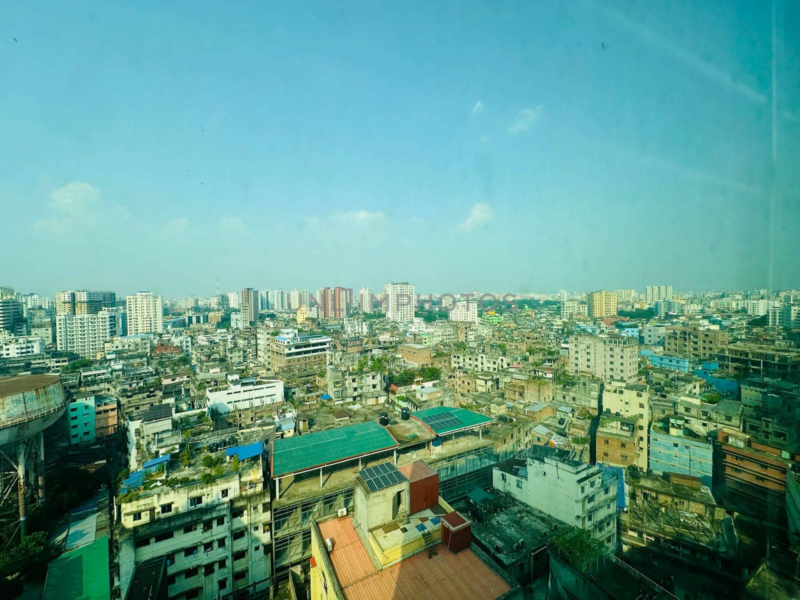 A panoramic view of the cityscape of Dhaka, Bangladesh, on a sunny day. The image shows a dense urban environment with buildings of varying heights. The sky is clear and blue, with a few white clouds. The buildings are mostly brown and gray, with some green vegetation visible.