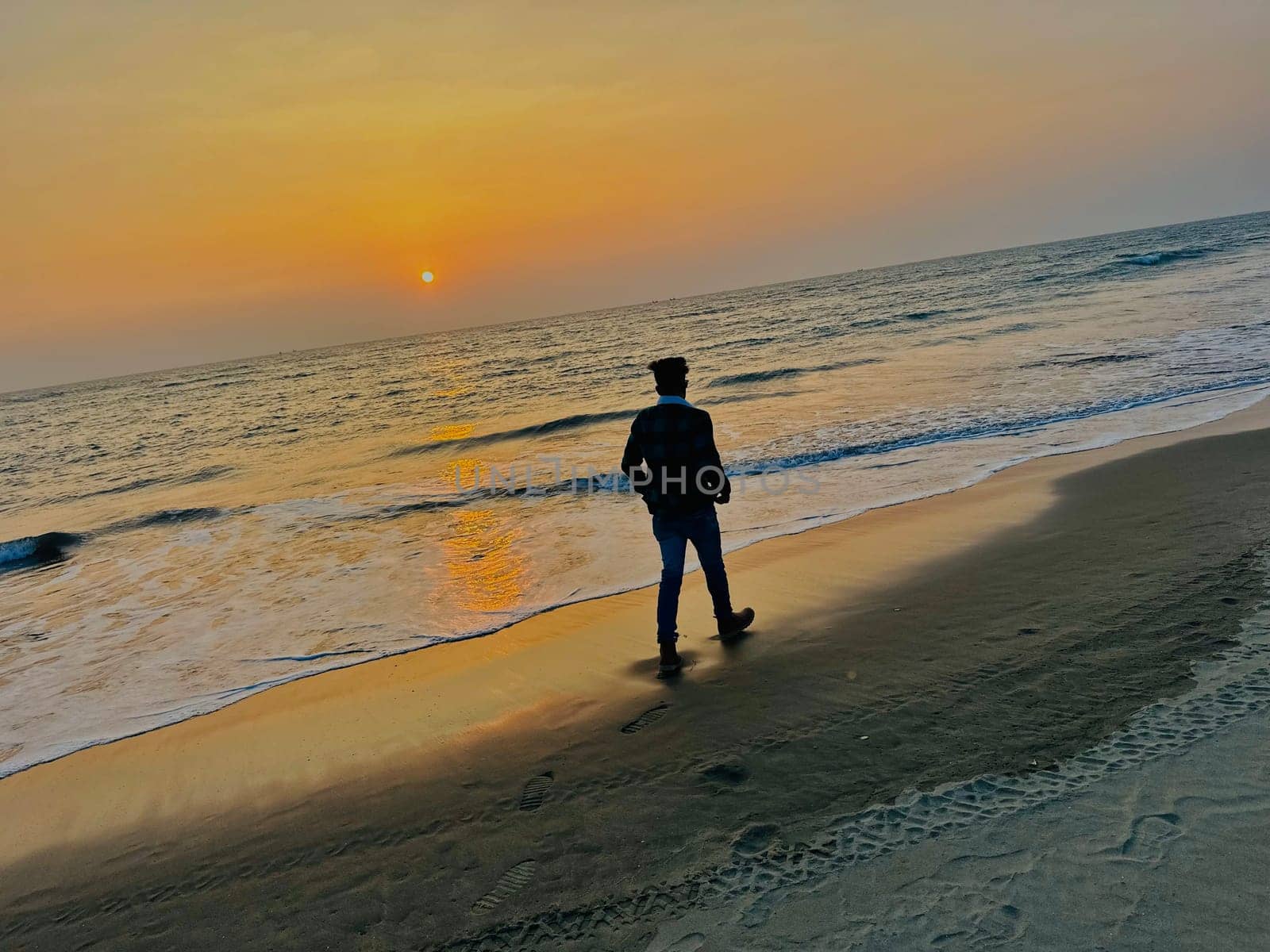 A man walks along a sandy beach towards the ocean as the sun sets, casting a golden reflection in the water. The sky is a warm orange and the water is a light blue.