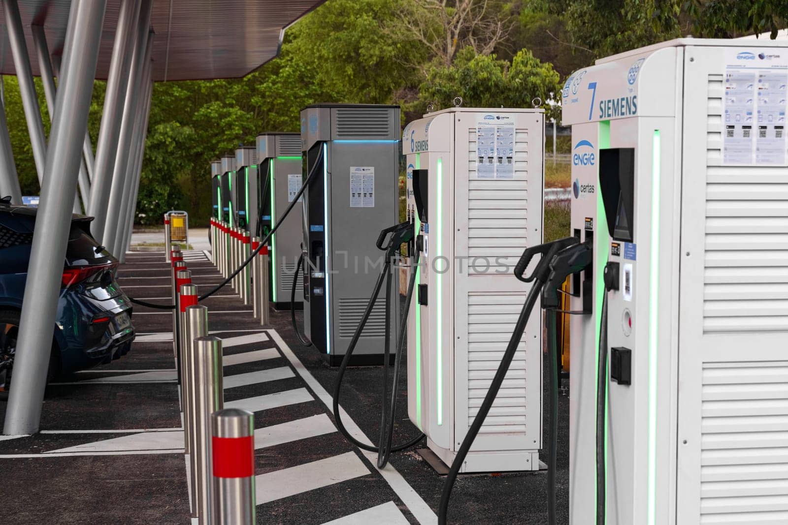 La Turbie, France - June 12, 2023: Multiple electric vehicle charging units lined up in an outdoor parking area, with cables connected to a parked car, indicating active charging.