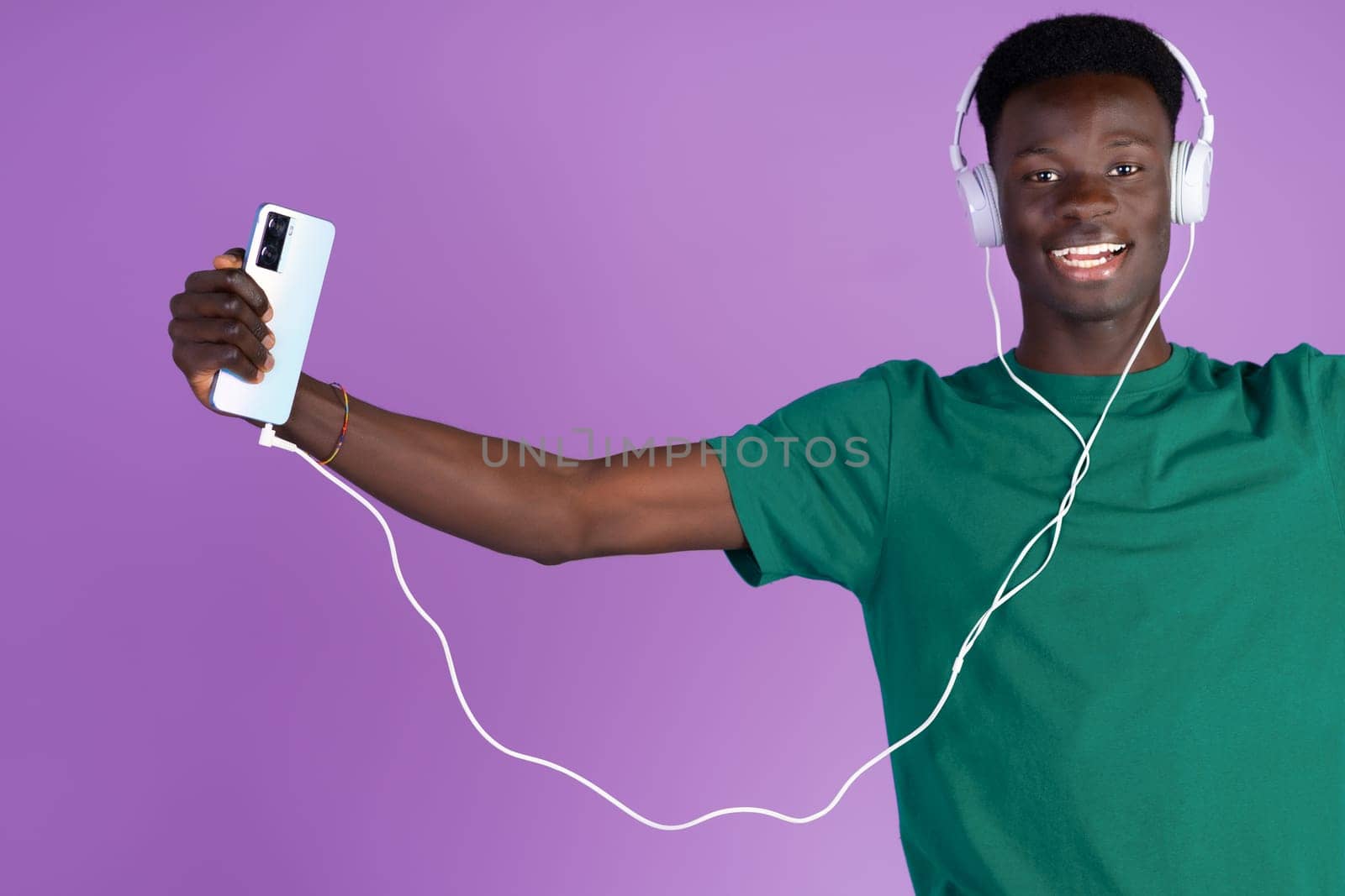 Happy young African man listening to music and dancing with headphones connected to his phone with a lilac background. by Ceballos