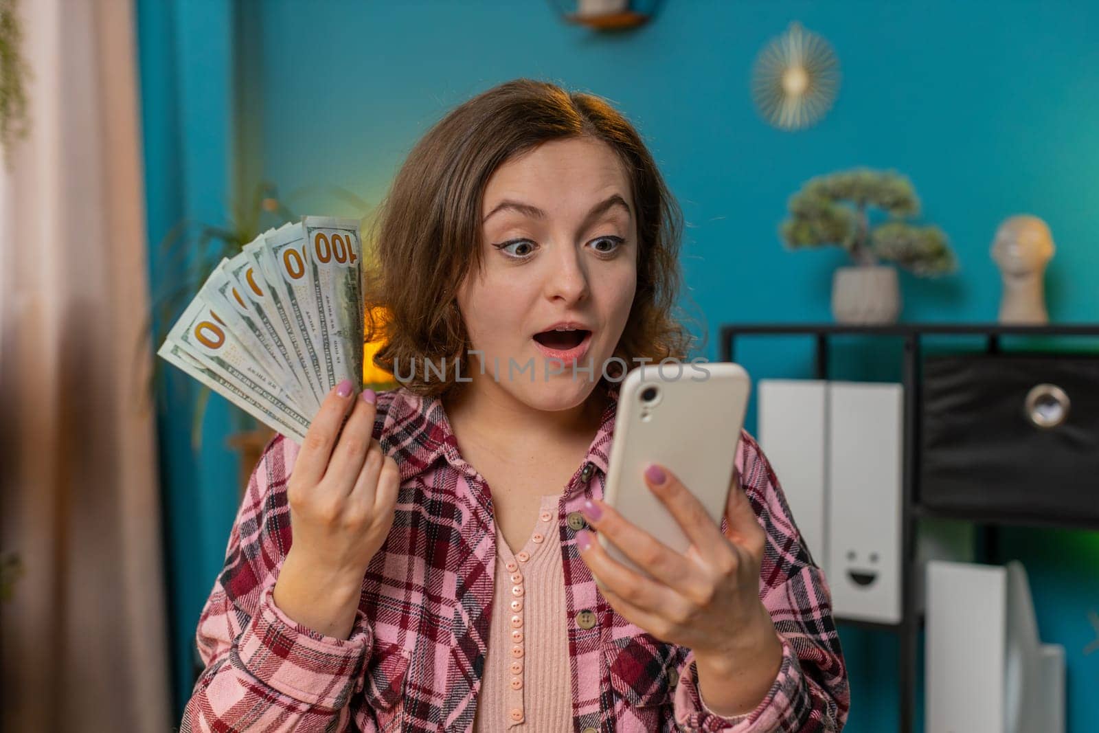 Rich excited amazed shocked young woman freelancer looks at camera holding money cash and smartphone. Female businesswoman girl satisfied of income earnings saves money sitting at home office at table