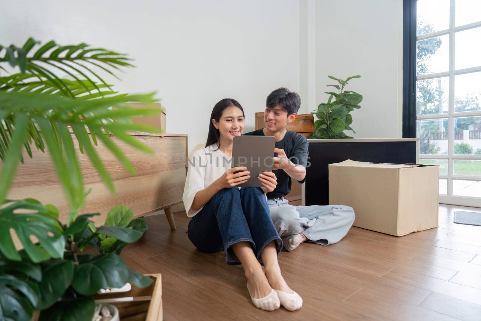 A cheerful couple sitting on the floor of their new home, surrounded by moving boxes and plants, using a tablet to plan their interior design.