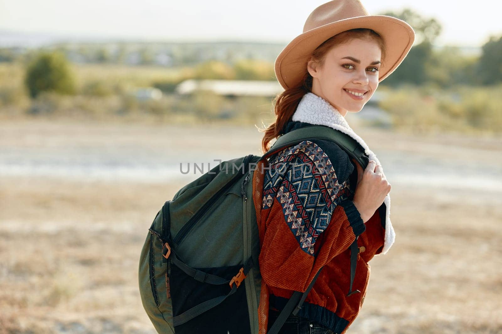 Woman in stylish hat and cozy sweater standing in field with backpack on a sunny day