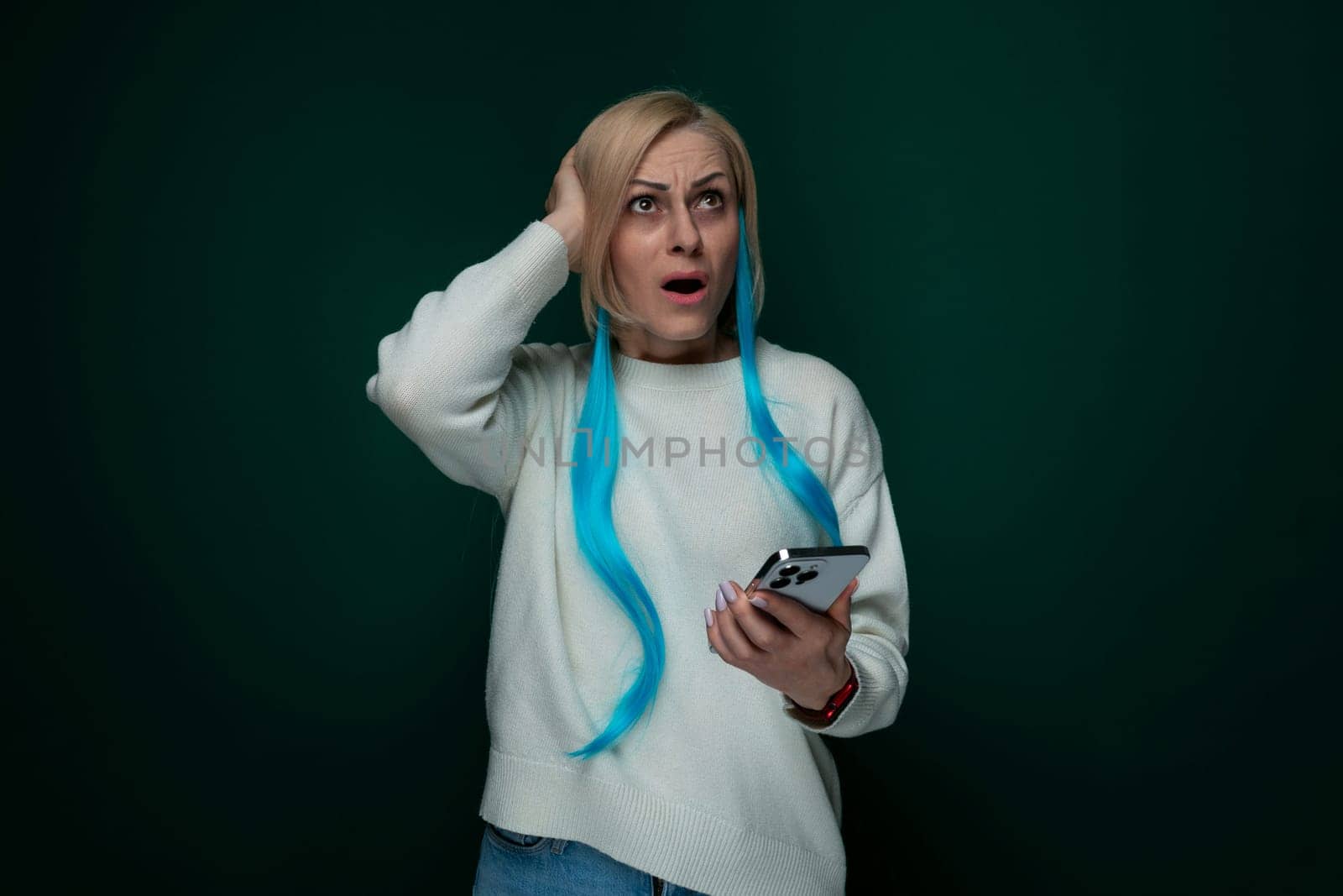 A woman with vibrant blue hair is seen holding and looking at a cell phone in her hand. She appears focused while interacting with the device, possibly texting or browsing. The background is blurred, emphasizing the main subject.