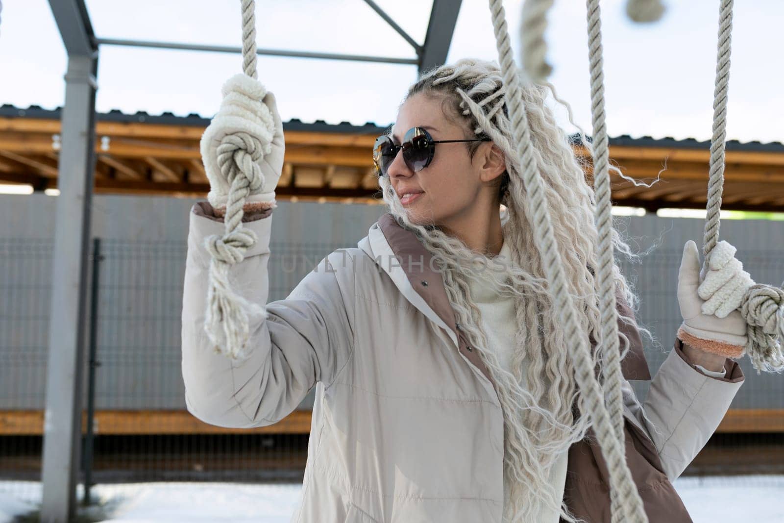 A woman wearing sunglasses and a scarf is seated on a swing, gently swaying back and forth. The swing is suspended from a sturdy frame, and the womans expression is relaxed.