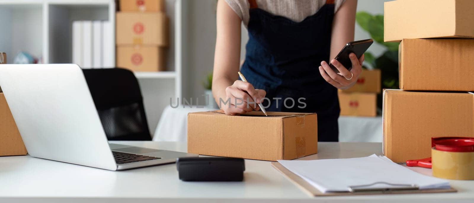 A small business owner prepares a parcel box for delivery, using a laptop and smartphone in an online ecommerce store setting.