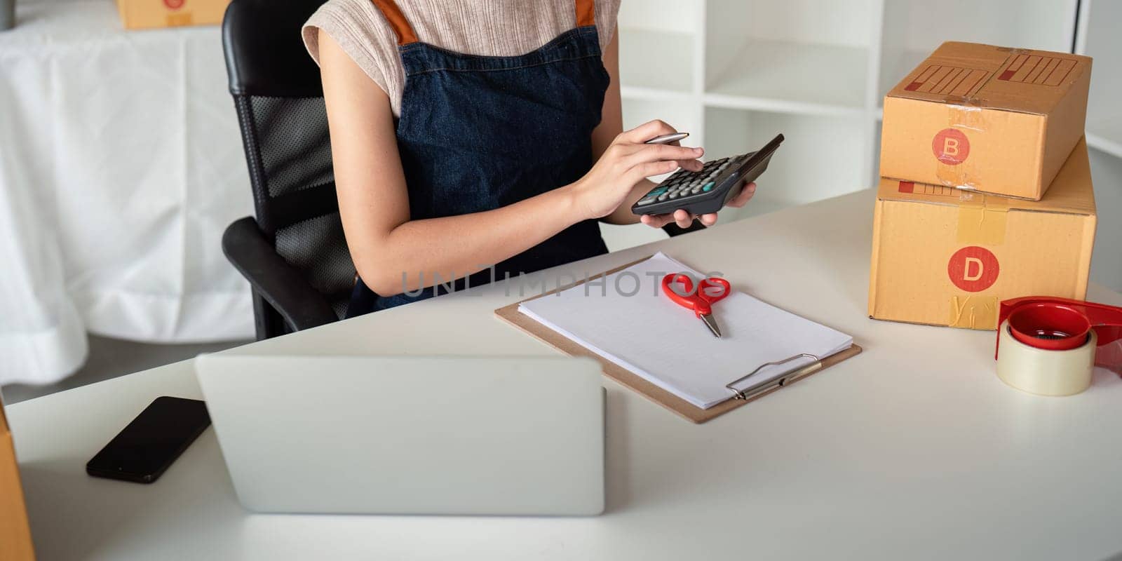 A young entrepreneur prepares parcel boxes for delivery, using a laptop and calculator, symbolizing the modern online ecommerce business environment.