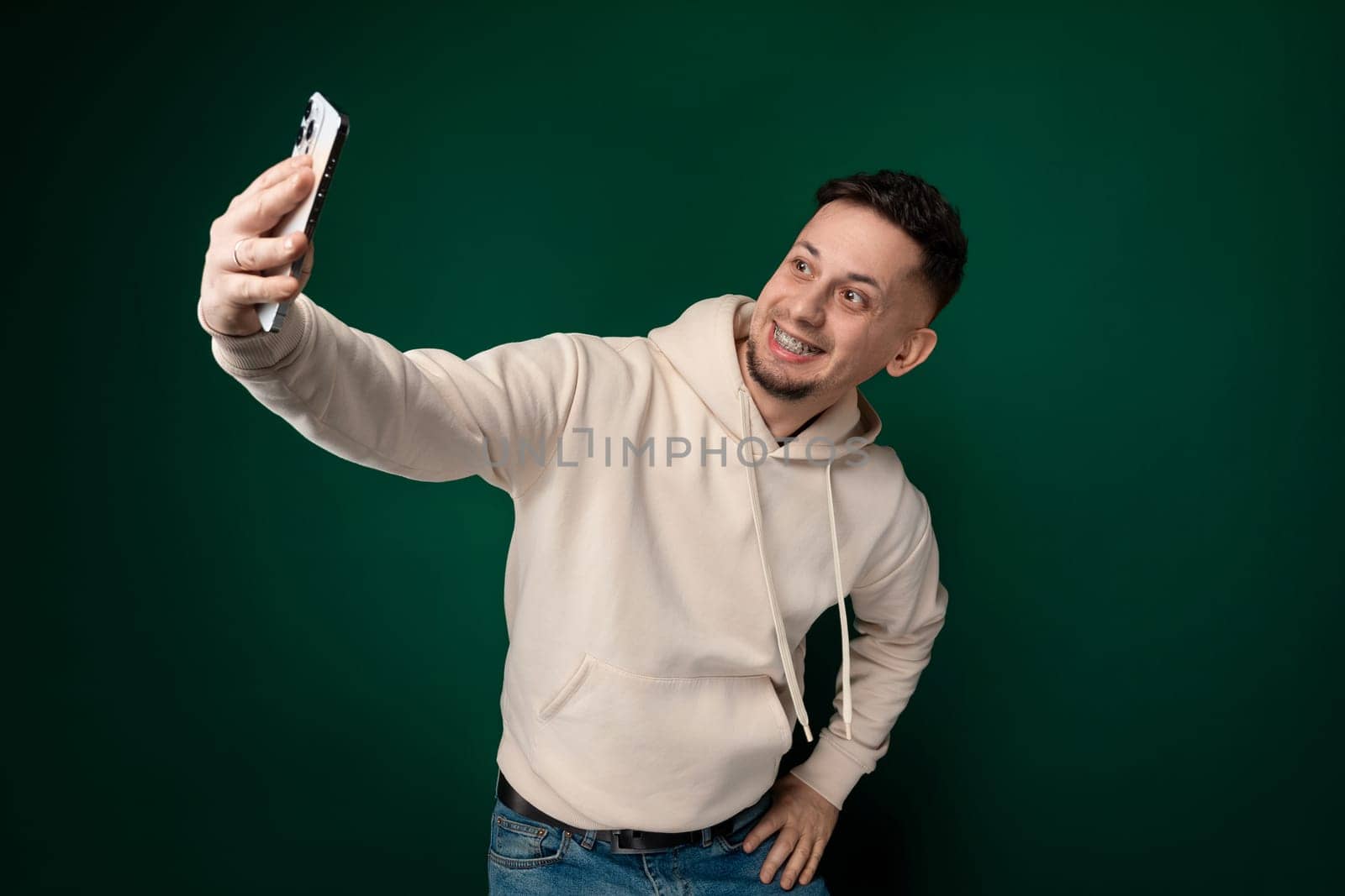 A man holding a cell phone at arms length, smiling and posing for a selfie. The background shows a city street with buildings and people passing by.