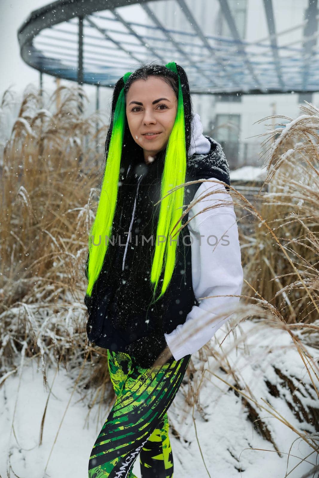A woman with vibrant green hair stands in a snowy landscape, the cold white snow contrasting with her colorful hair. She looks around, her breath visible in the chilly winter air.