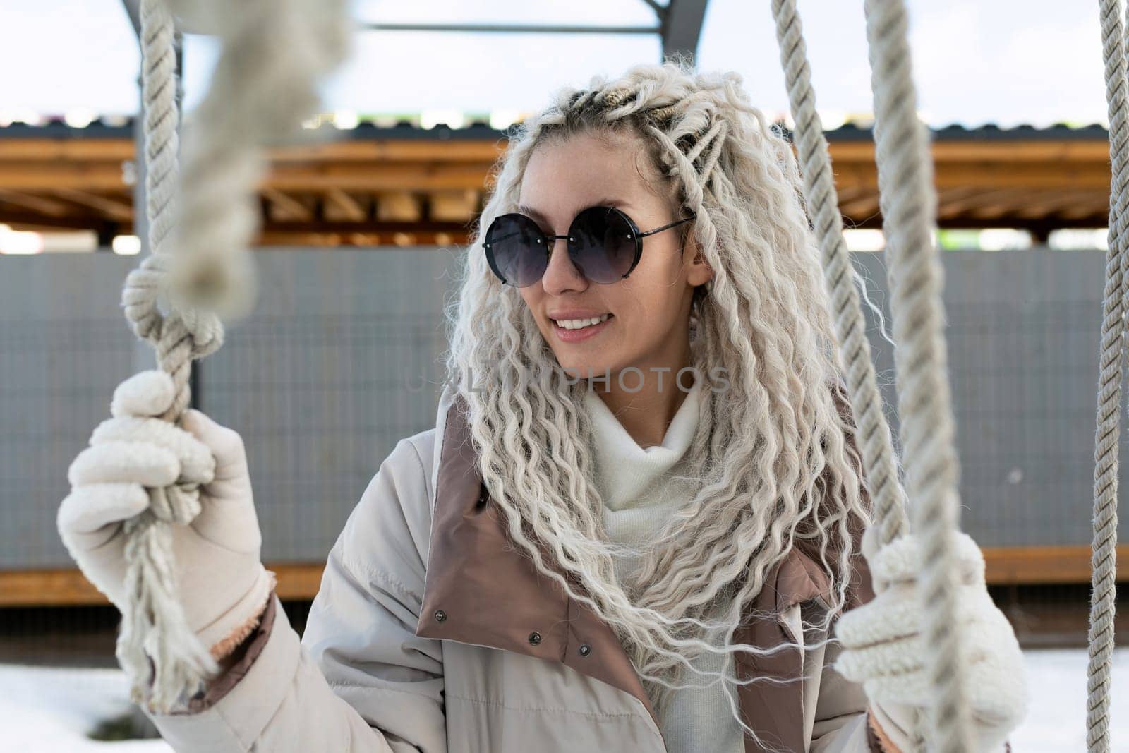 A woman is sitting on a swing, wearing sunglasses and a scarf. She appears relaxed as she swings back and forth gently.