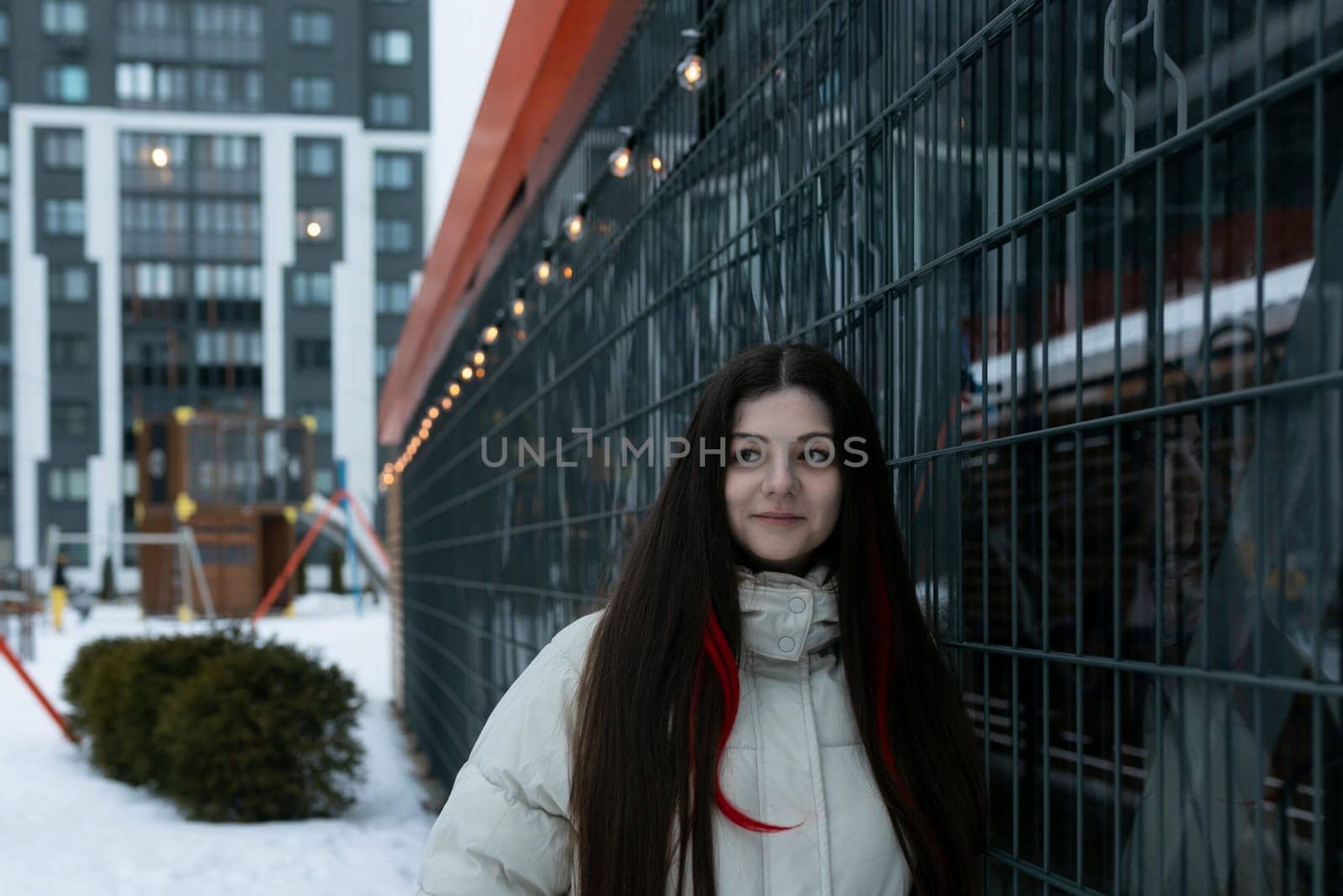 A woman is standing in front of a wooden fence covered in snow. The scene is simple, with the woman looking straight ahead, wearing warm clothing.