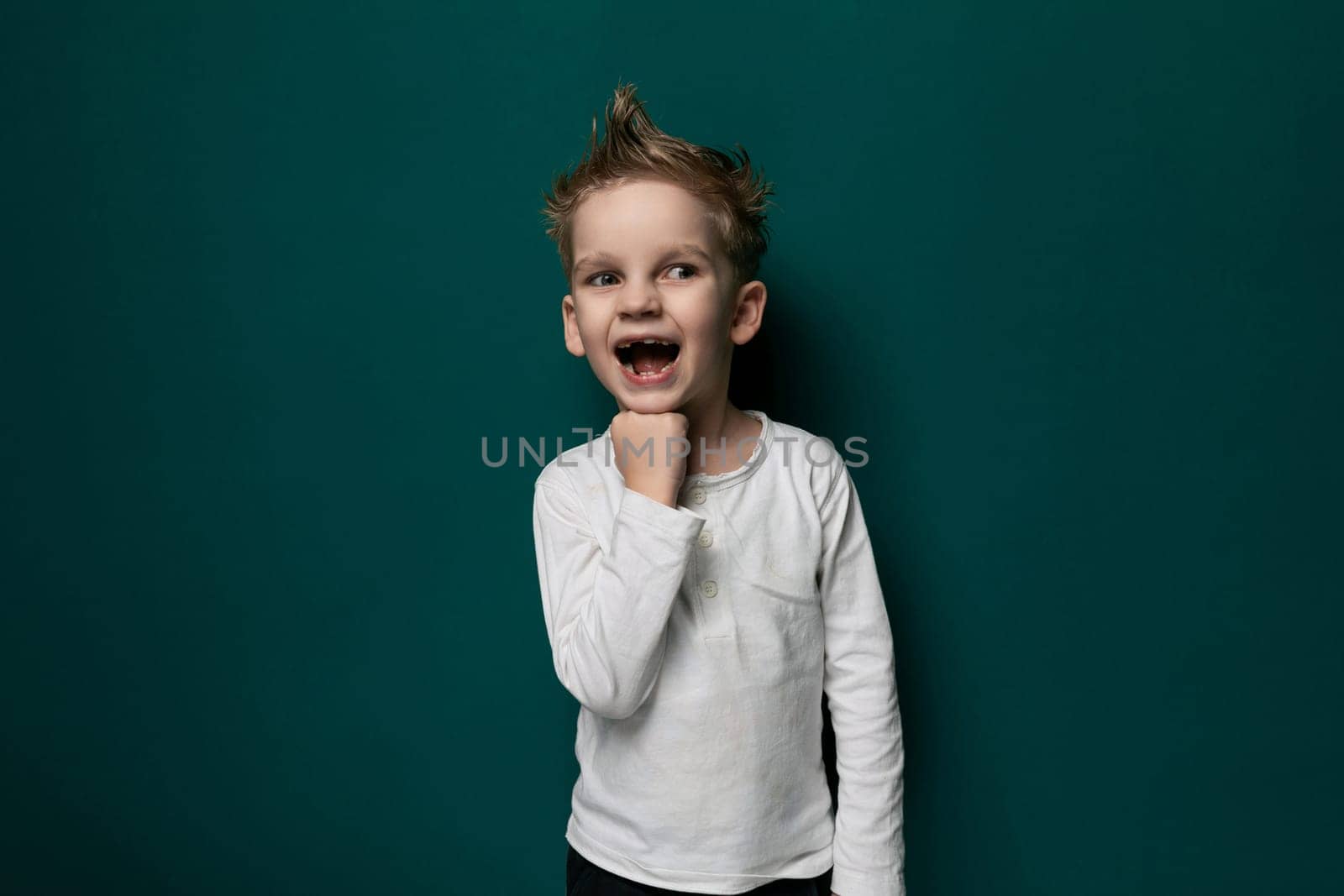 A young boy in casual clothing stands in front of a bright green wall, looking straight ahead with a neutral expression. The wall serves as a simple backdrop to the childs presence in the scene.