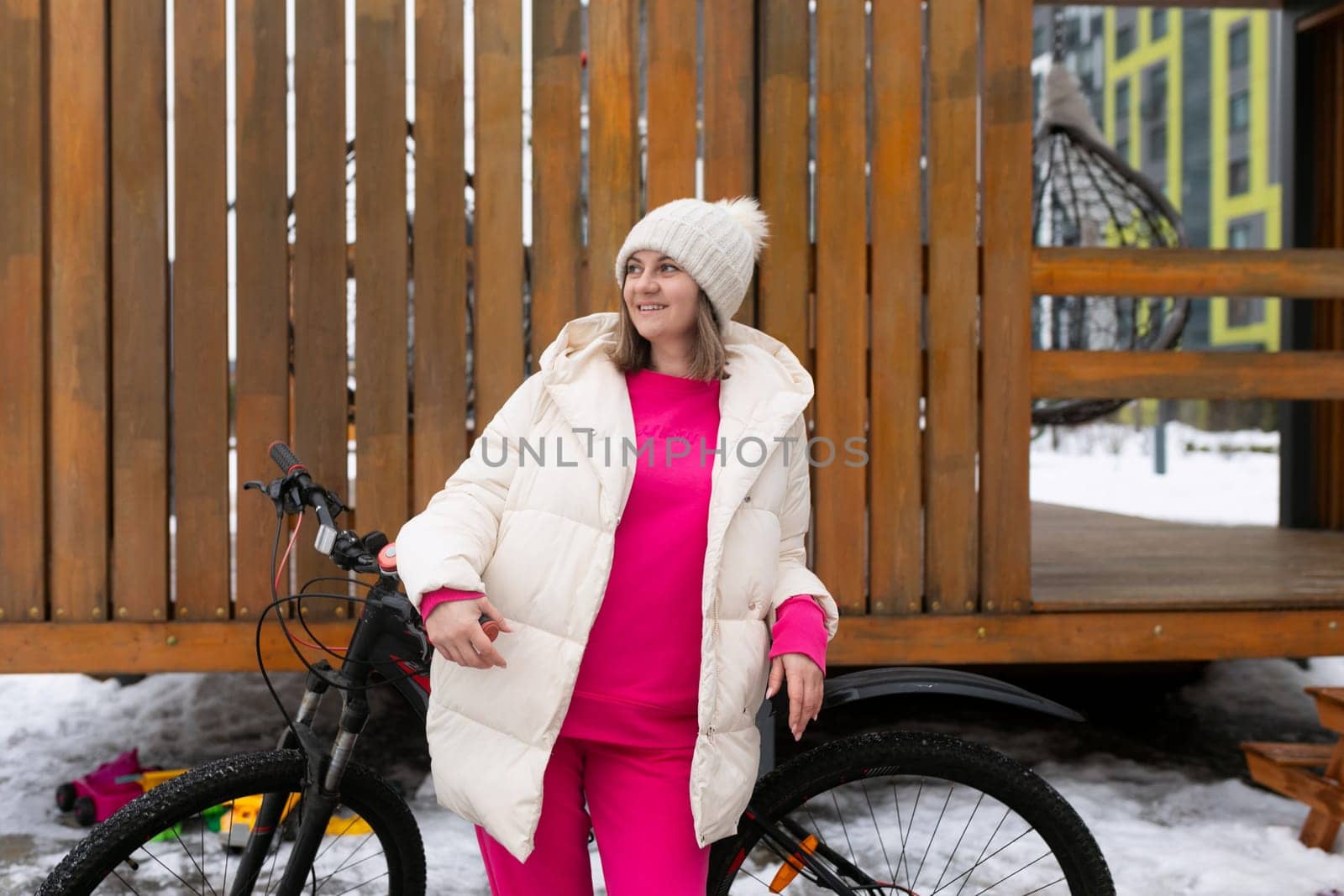 A woman stands next to a bicycle in a snowy landscape, surrounded by white fluffy snow. She is wearing warm winter clothing, looking at the bike.
