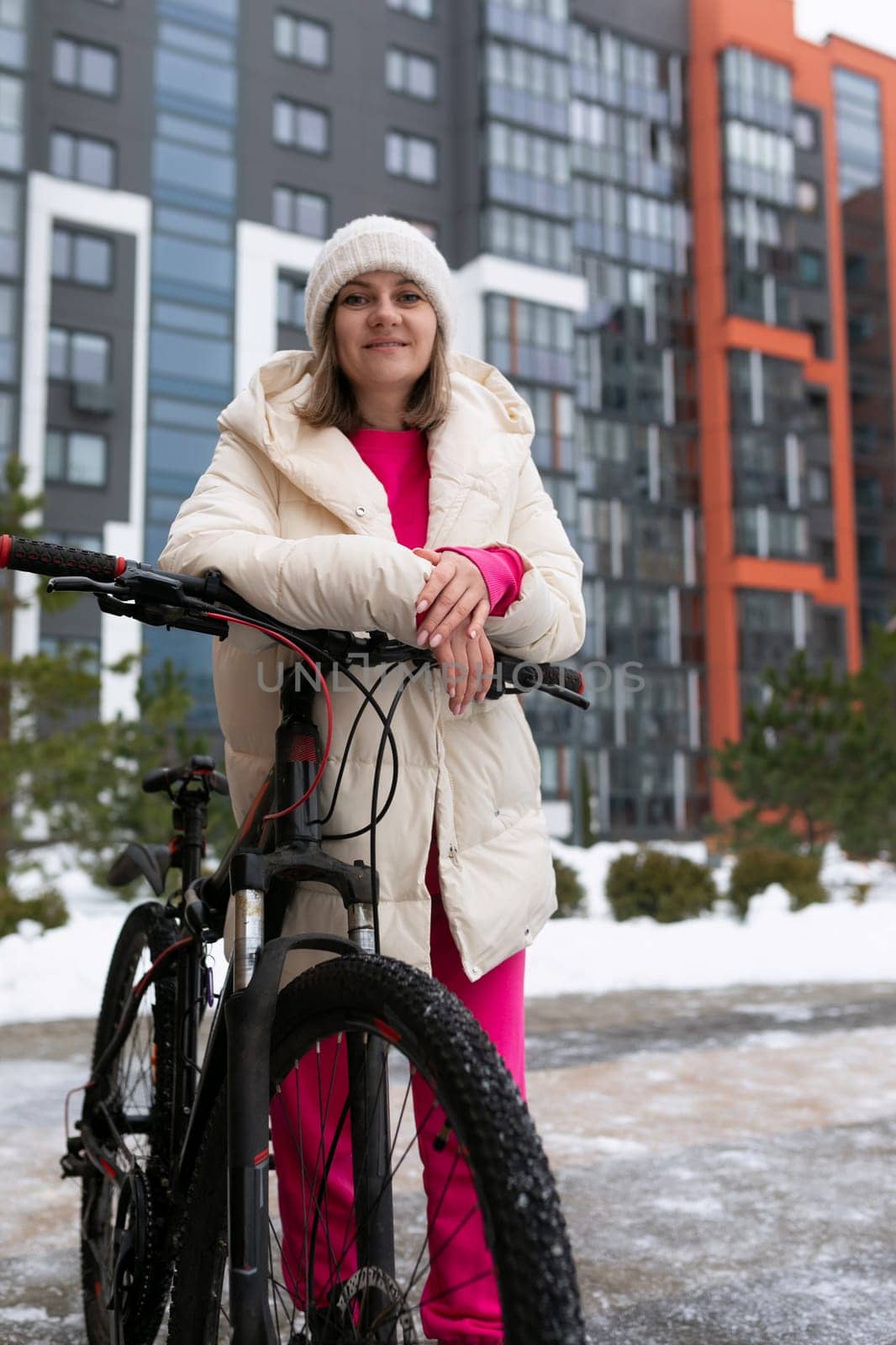 A woman stands next to a bike on a snowy day, wearing winter clothing. The snowy landscape is visible in the background, showcasing a cold and wintery scene.