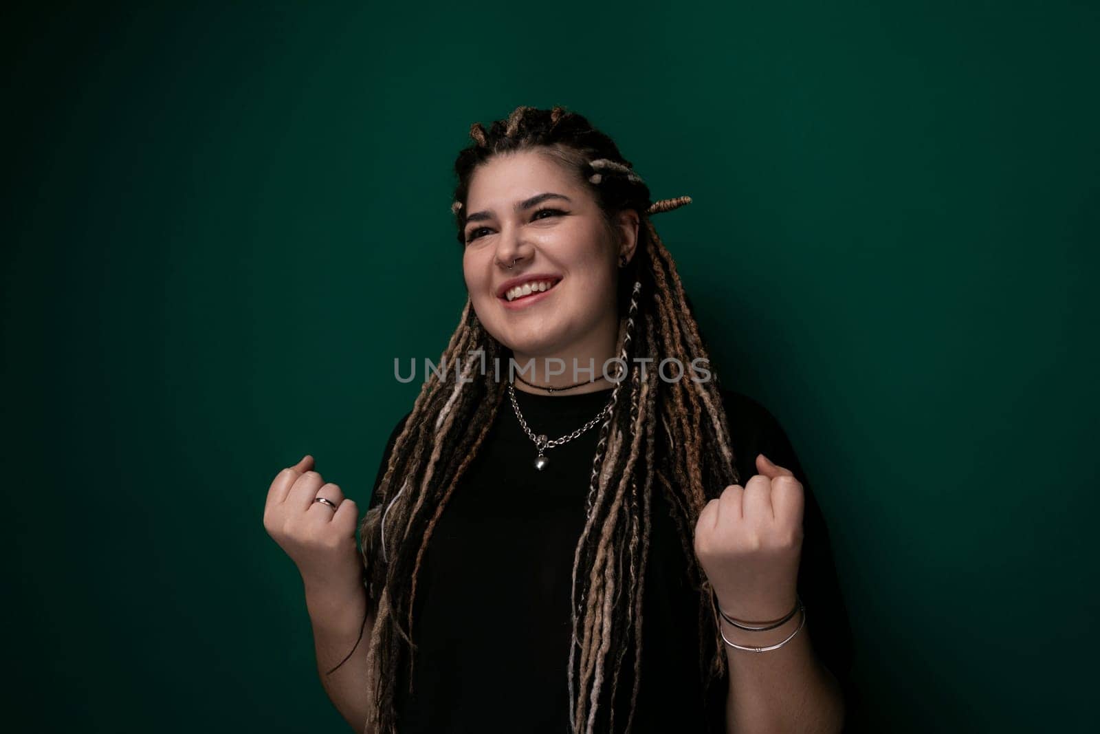A woman with dreadlocks is standing in front of a bright green wall. She has a confident posture and is looking directly at the camera. The contrast between her hair and the wall creates a striking image.