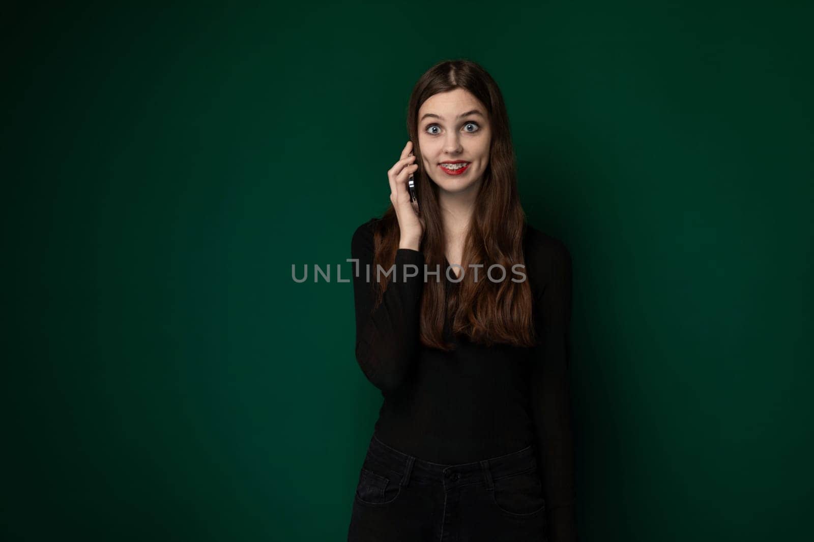 A woman standing in front of a green wall, holding a cell phone to her ear and engaged in conversation. She appears focused on the call, with a neutral expression on her face.
