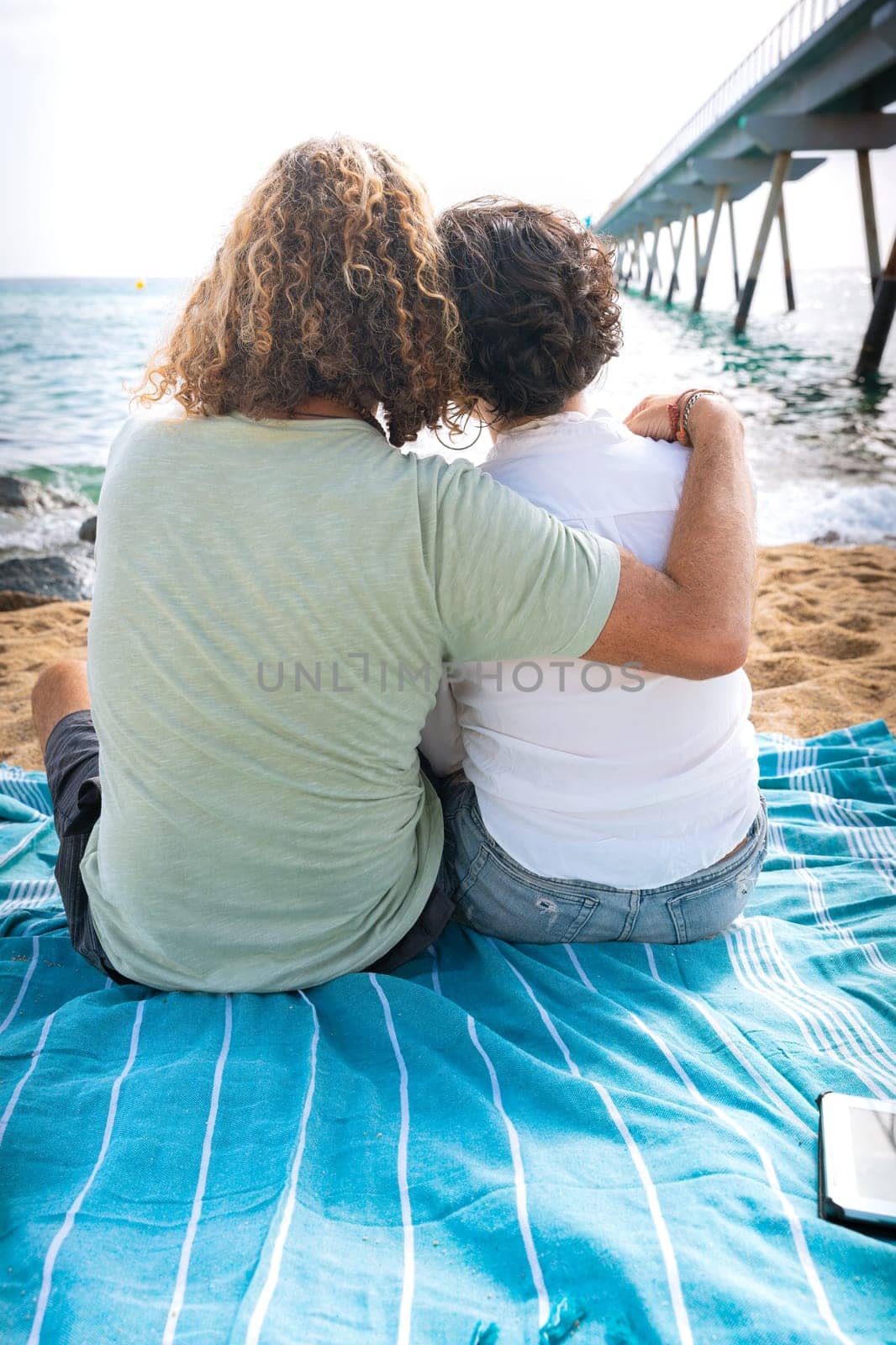 Rear view two happy friends sitting on the beach on vacation,looking at the sea. by mariaphoto3