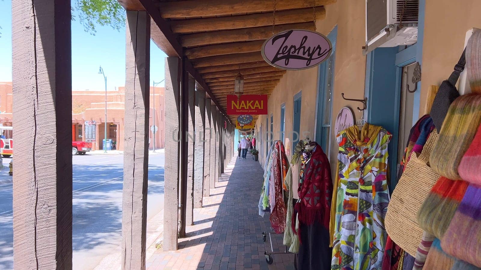 Santa Fe, New Mexico, USA-June 11, 2024-Slow motion-A picturesque covered walkway in Santa Fe, New Mexico, featuring traditional adobe architecture and vibrant blue window frames. The pathway is lined with shops, including a silversmith, showcasing the city rich cultural heritage and lively atmosphere. Potted plants add a touch of greenery to this charming scene.