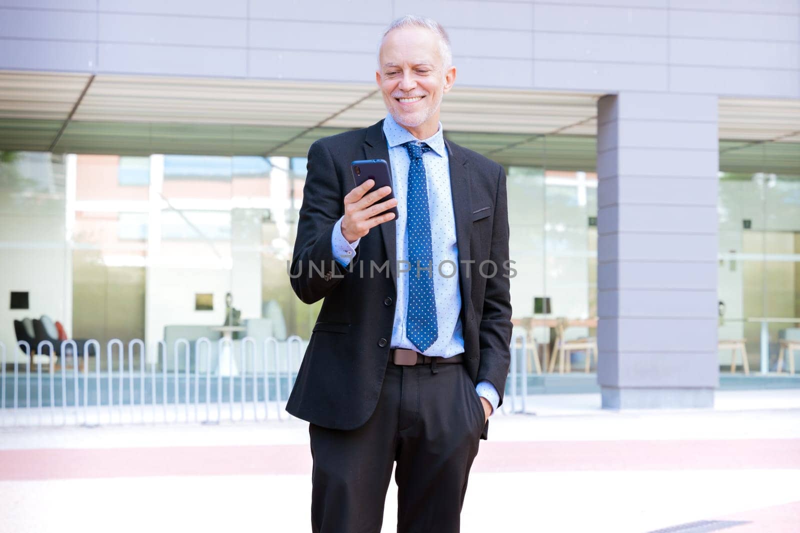 Satisfied with the results, the businessman walks on the street outside the office building, a mature boss holds a phone in his hands, writes messages and reads news online, using an application.