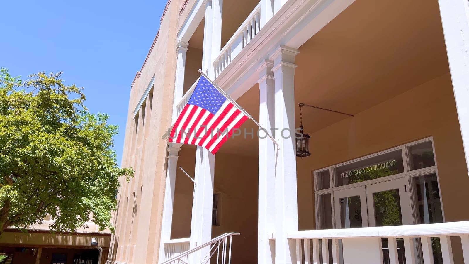 Santa Fe, New Mexico, USA-June 10, 2024-Slow motion-A historic building in a downtown area featuring a symmetrical facade with two levels of balconies, an American flag, and well-maintained landscaping on a sunny day.