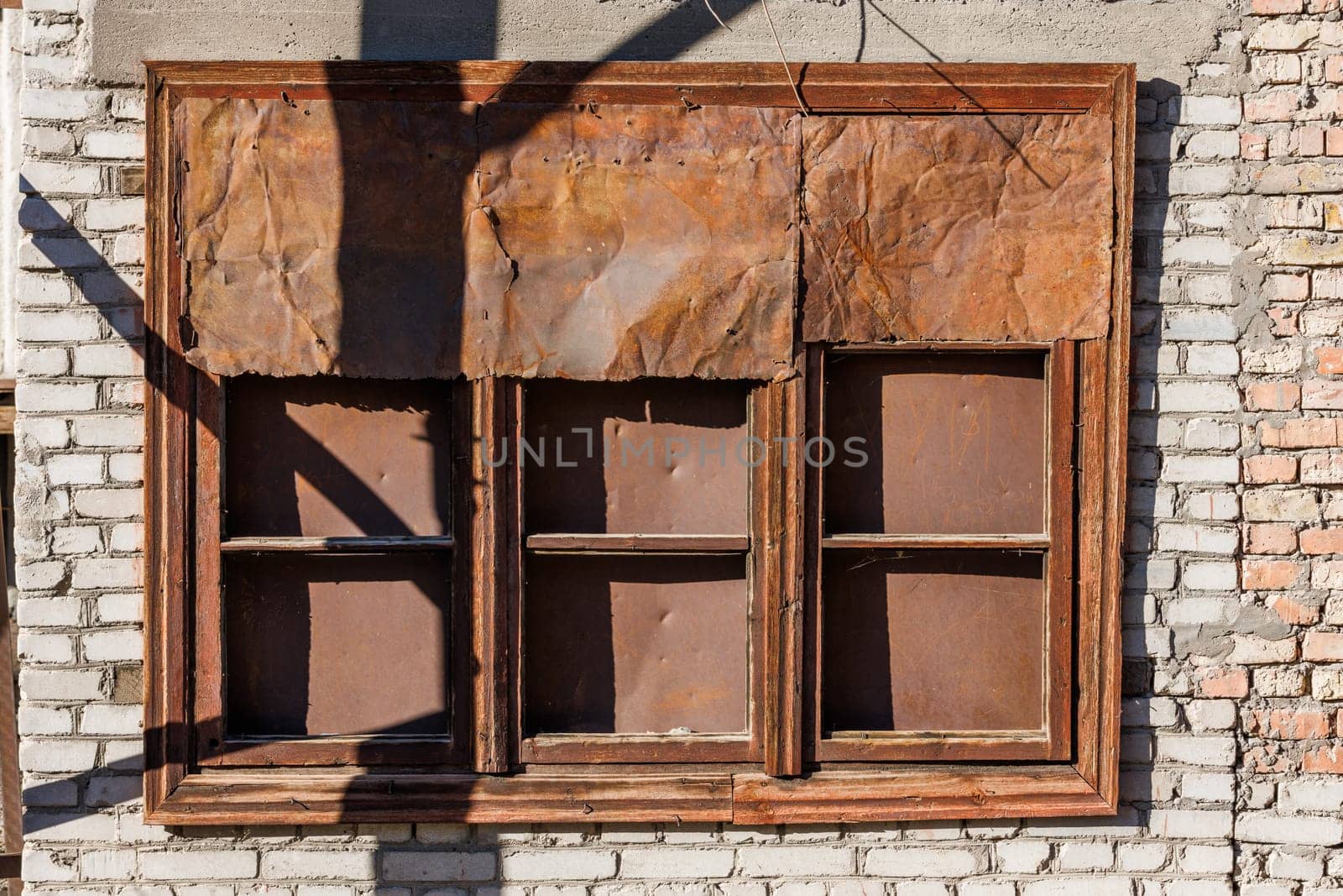 old brown three-sectioned wooden window covered with rusted sheet metal on white brick wall under direct sun light.