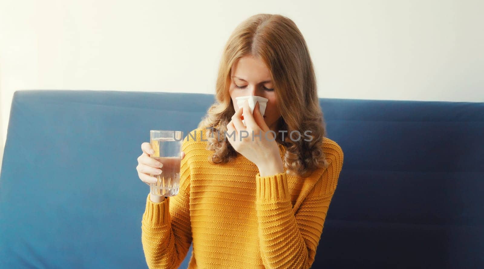 Sick exhausted young woman sneezing blow nose using tissue drinks water from a glass taking a medication at home