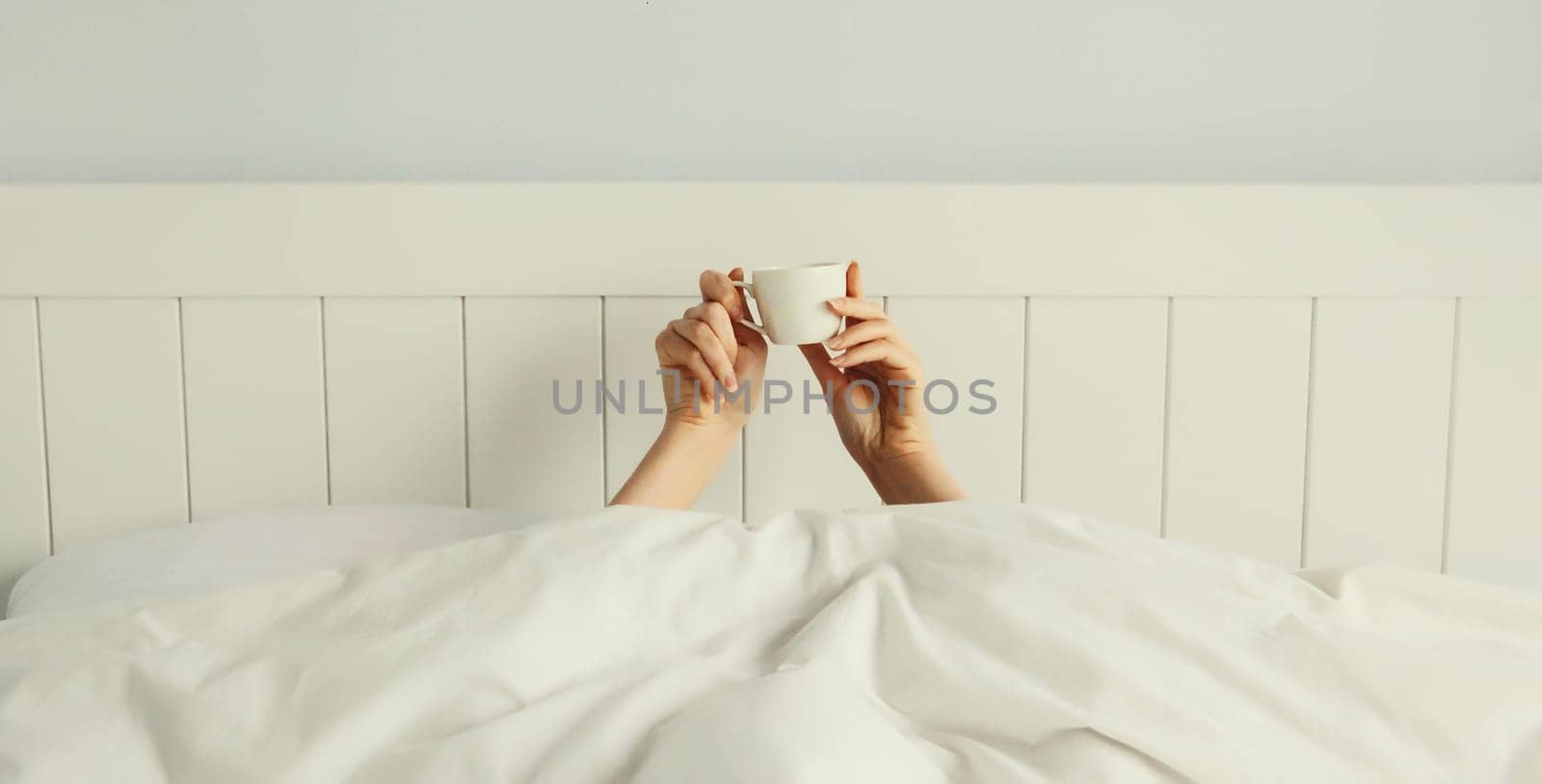 Cheerful lazy woman waking up after sleeping lying in soft comfortable bed showing empty cup coffee stretching her hands up from under the blanket in white bedroom
