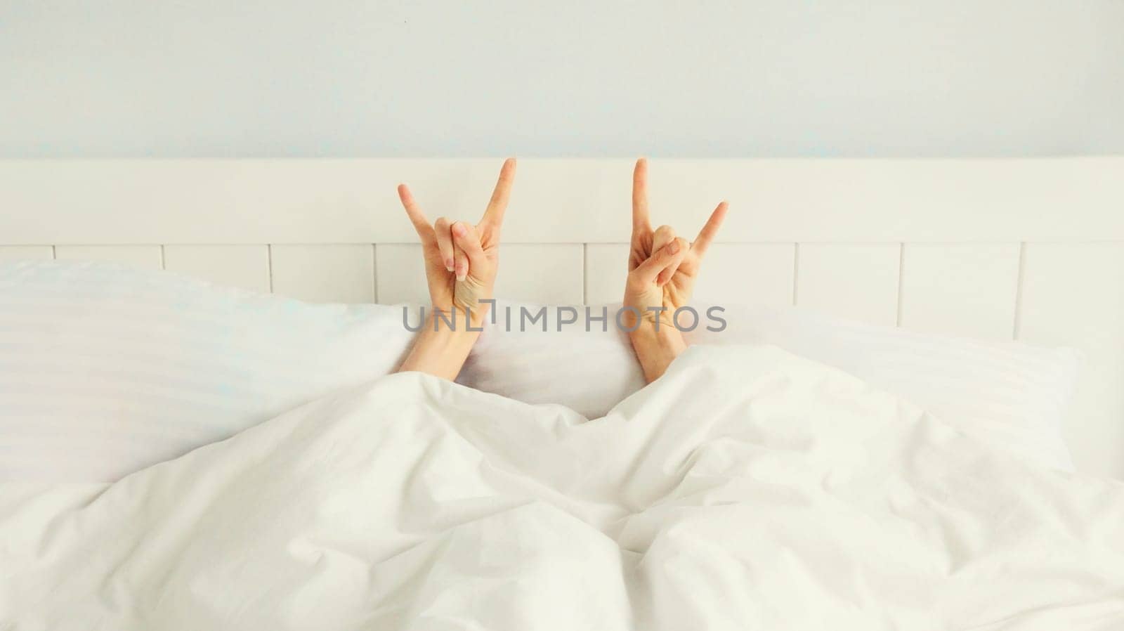 Cheerful lazy woman waking up after sleeping lying in soft comfortable bed showing gesture stretching her hands up from under the blanket in white bedroom