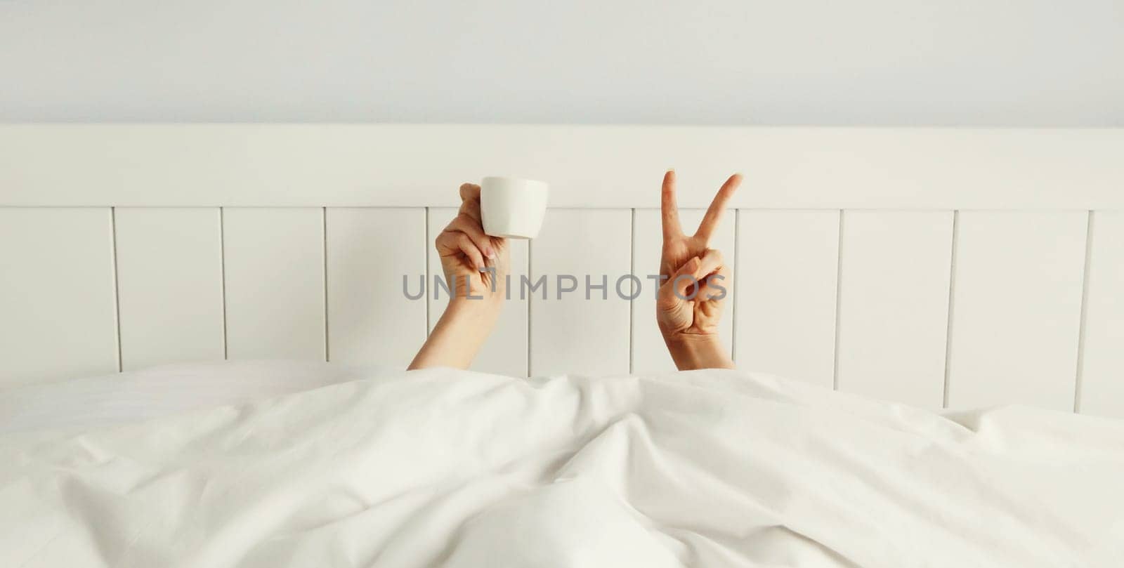 Cheerful lazy woman waking up after sleeping lying in soft comfortable bed showing empty cup coffee stretching her hands up from under the blanket in white bedroom