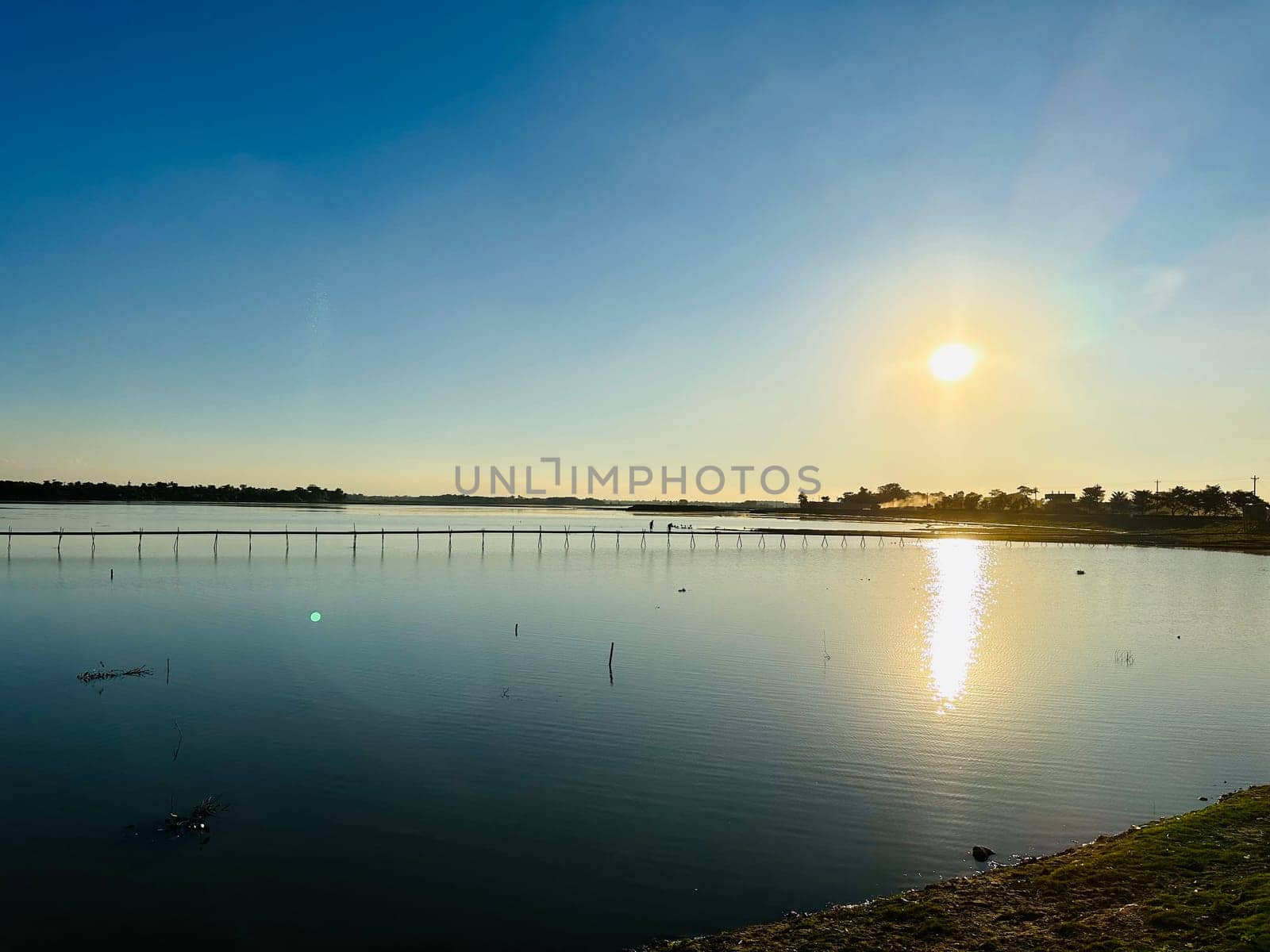 A peaceful sunset over a serene lake, with a long wooden bridge stretching across the water. The sun casts a warm glow on the rippling surface, creating a mesmerizing reflection. The sky is a vibrant blue, gradually fading into a soft orange hue as the sun dips below the horizon. The trees lining the shore are silhouetted against the fading light, adding a touch of mystery to the scene.