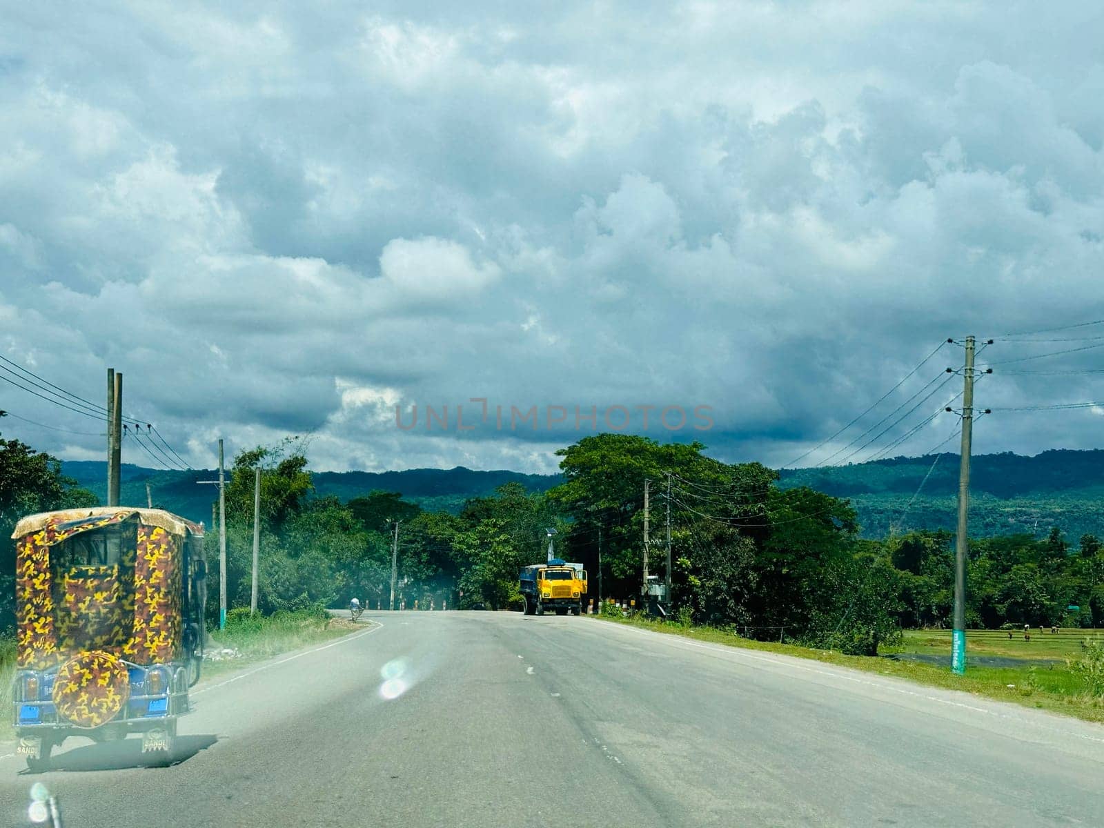This image shows a yellow truck driving down a winding road in rural Bangladesh. The road is surrounded by green trees and hills, and the sky is cloudy. The truck is the only vehicle on the road, and there are no people in sight.