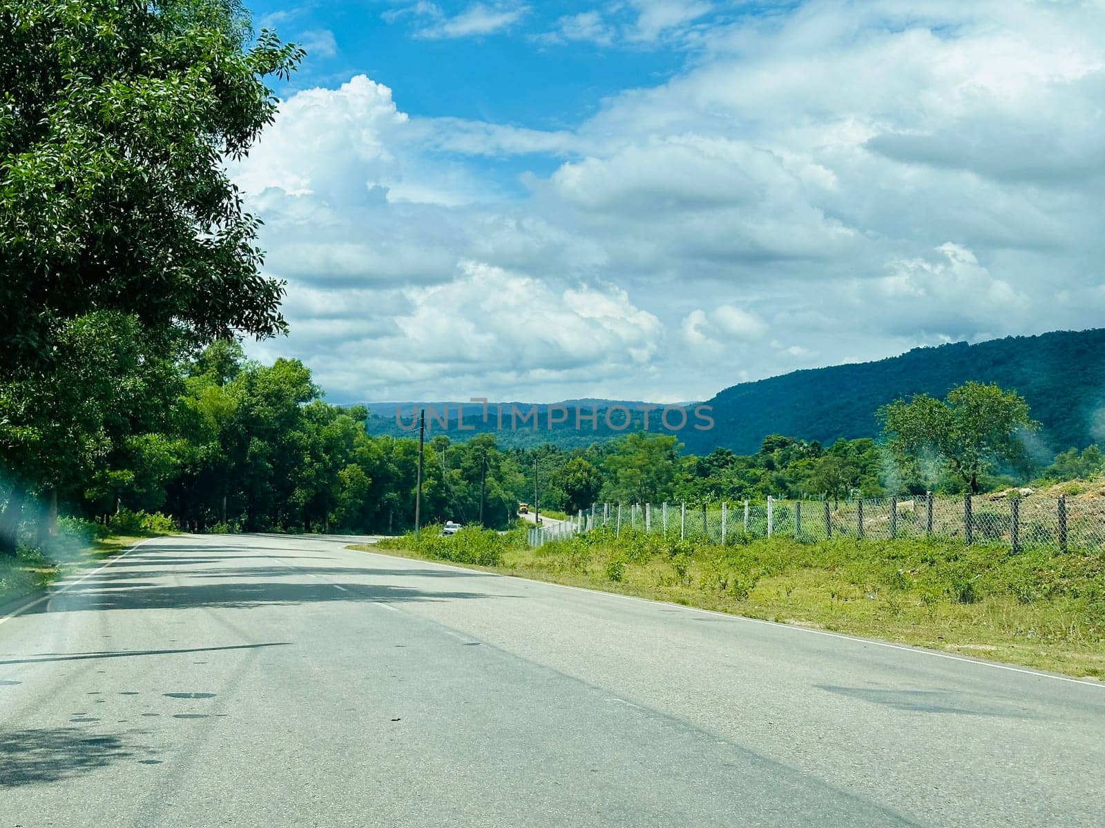 A scenic view of a winding asphalt road cutting through a lush green landscape, with dense trees lining the road on the left and a fence separating the road from a grassy field on the right. The road leads toward a distant mountain range under a clear blue sky dotted with white fluffy clouds.
