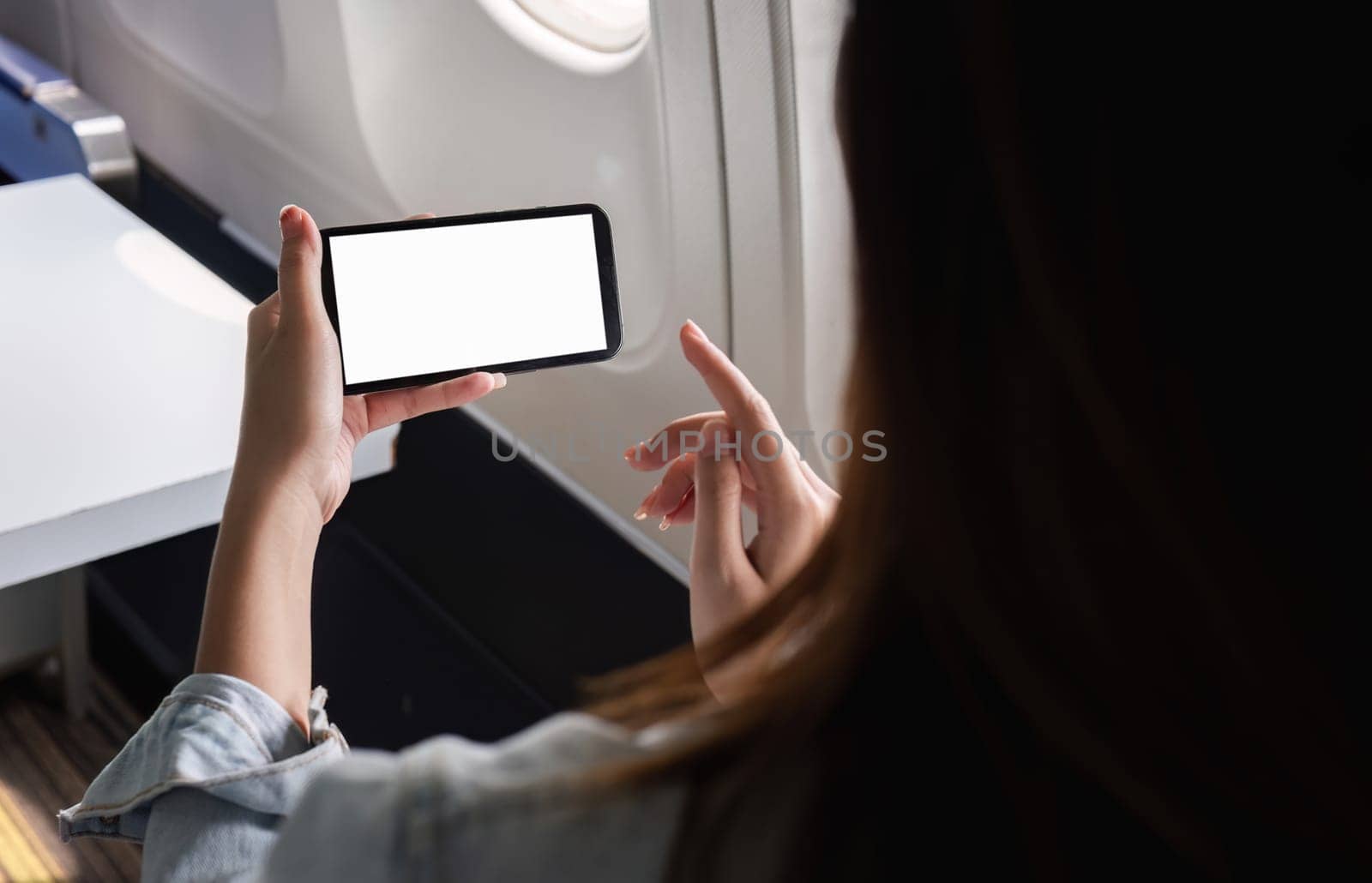 Young Businesswoman Using Smartphone While Traveling by Plane for Work, Focused on Blank Screen, Modern Technology in Business Travel by wichayada