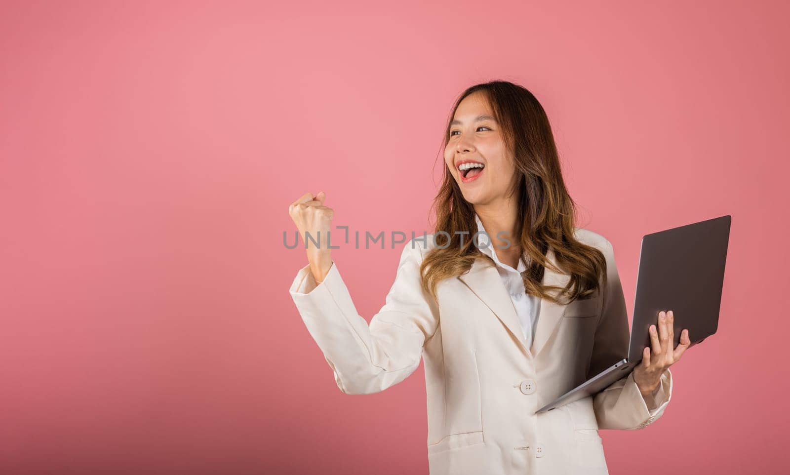 Portrait of happy Asian beautiful business woman teen confident smiling face holding using laptop computer and excited celebrating success, studio shot isolated on pink background, with copy space