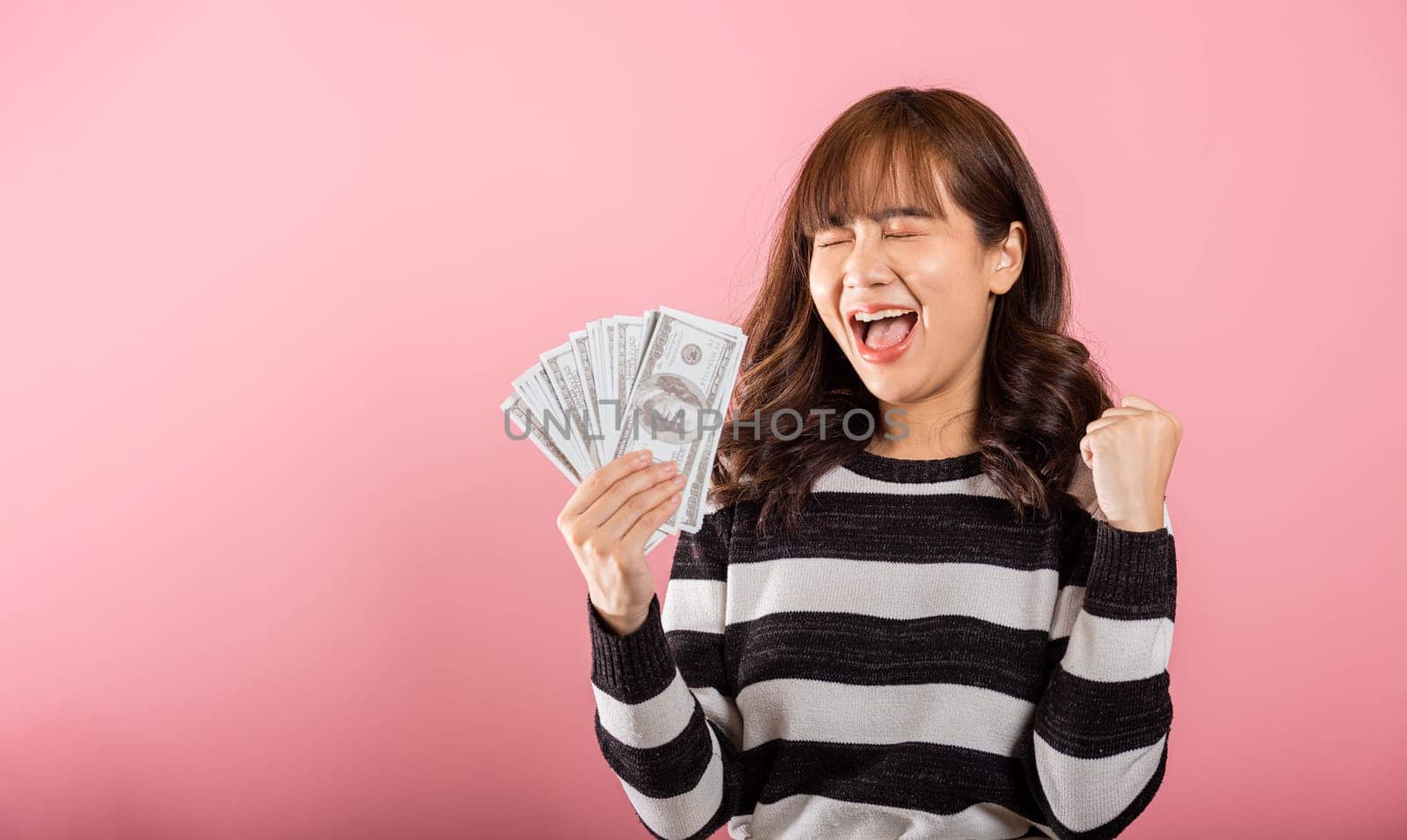 Asian happy portrait beautiful young woman standing smile her celebrating holding dollar money fan banknotes on hand and looking to camera on pink background with copy space for text