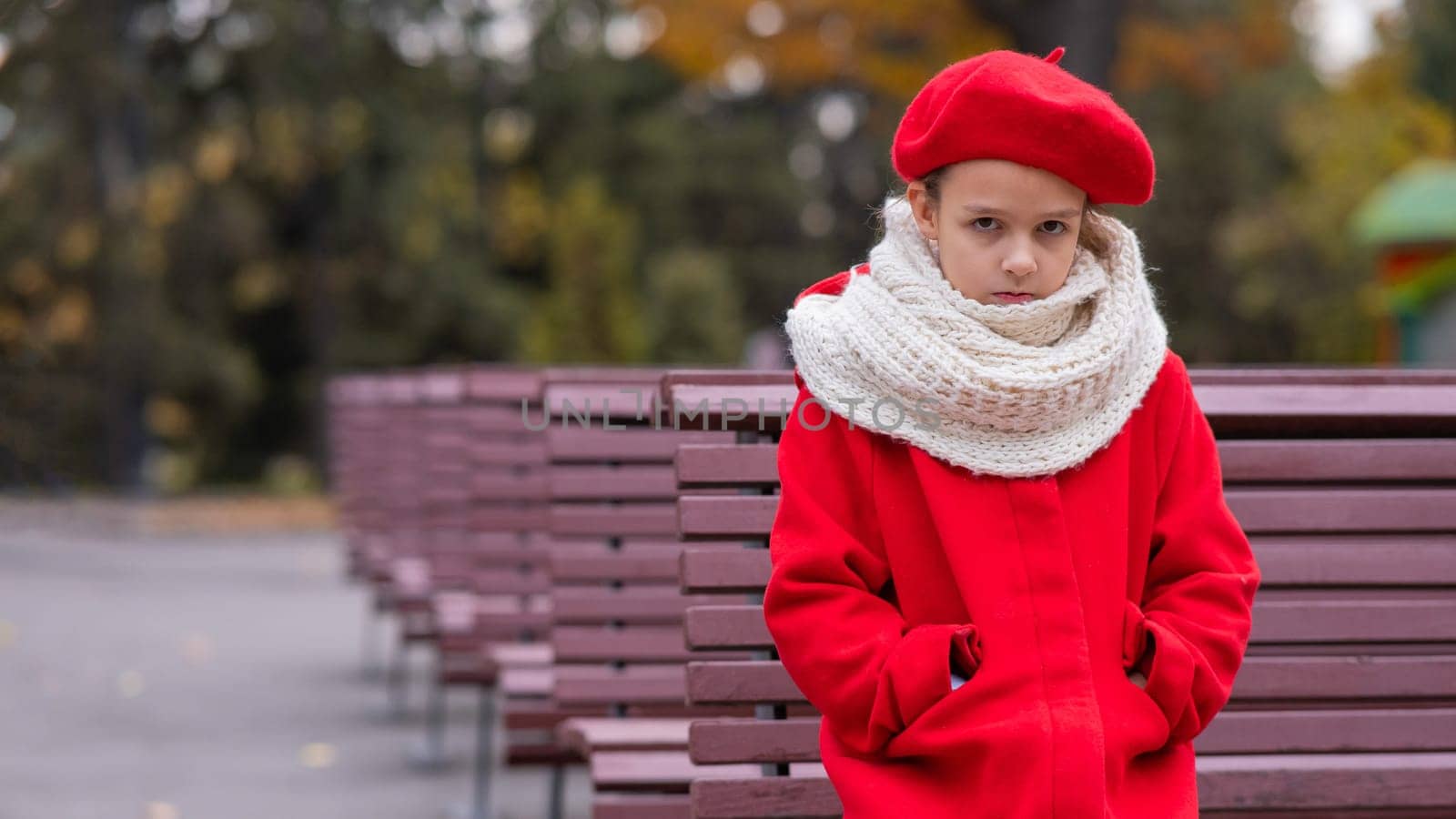 A sad Caucasian girl in a red coat and a beret sits alone on a bench