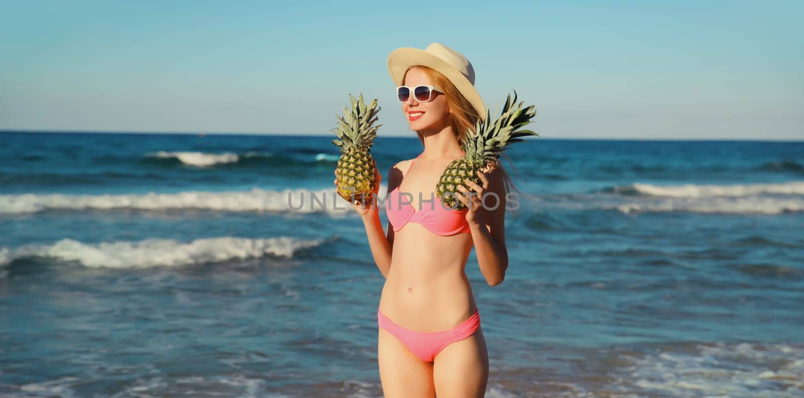 Summer vacation, beautiful happy young woman in pink bikini swimsuit, tourist straw hat holding pineapple fruits looking away on the beach on sea coast with waves