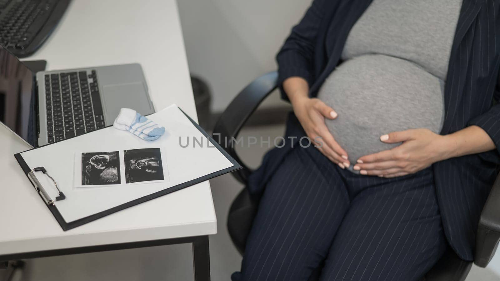 A pregnant woman works on a laptop in the office and looks at a photo from an ultrasound scan of the fetus. Belly close-up. by mrwed54