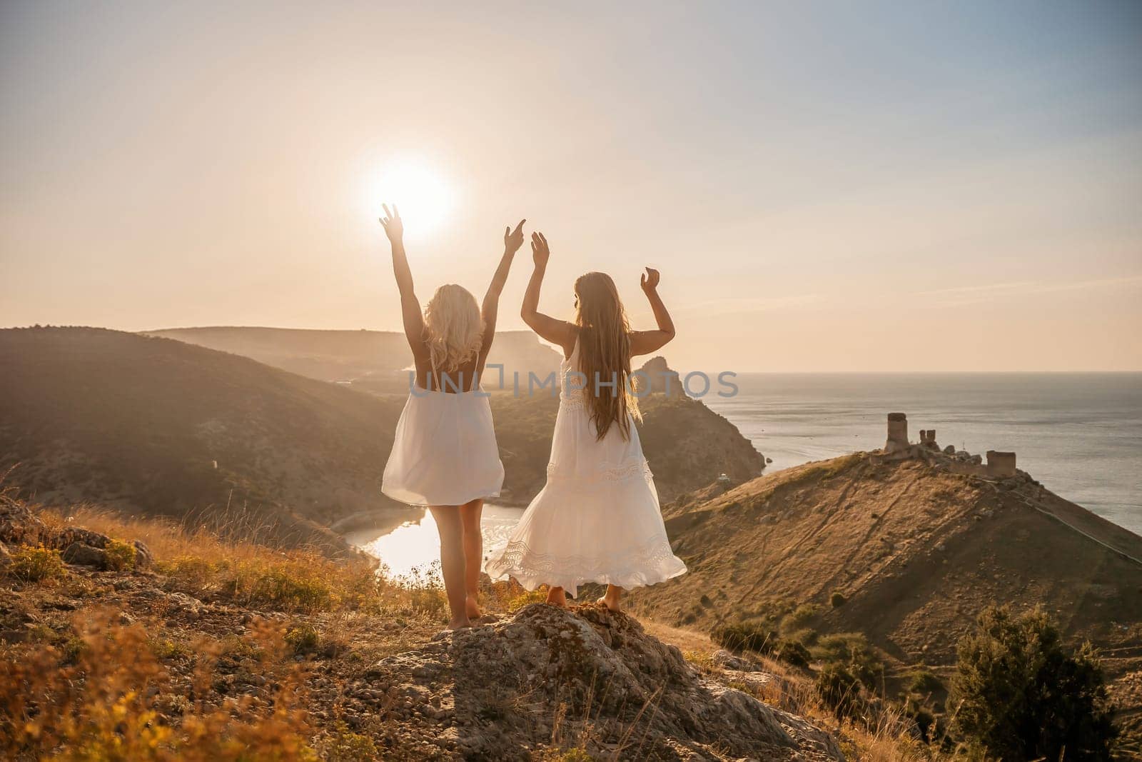 Two women are standing on a hill overlooking the ocean. They are holding hands and looking out at the water. The scene is peaceful and serene, with the sun shining brightly in the background. by Matiunina