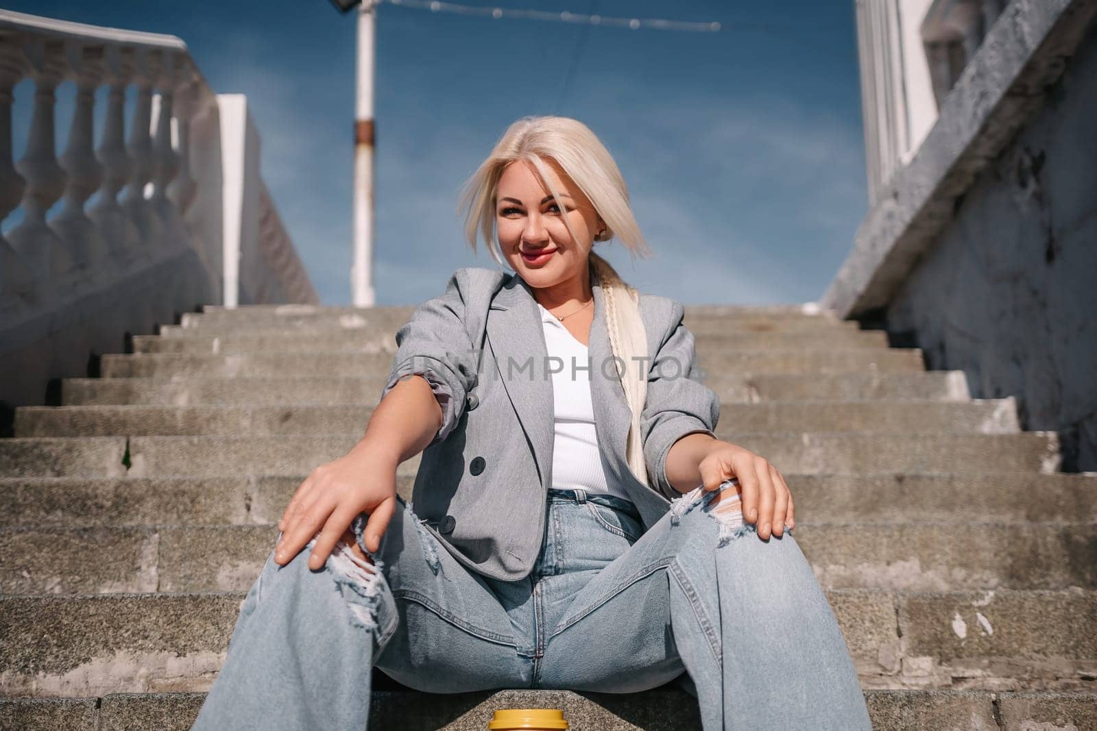 A woman in a gray jacket and blue jeans sits on a set of stairs. She is smiling and she is enjoying herself