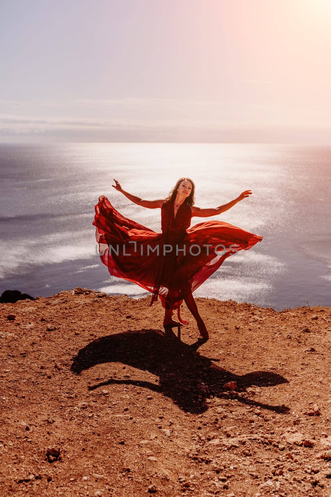 Woman red dress sea. Female dancer in a long red dress posing on a beach with rocks on sunny day. Girl on the nature on blue sky background