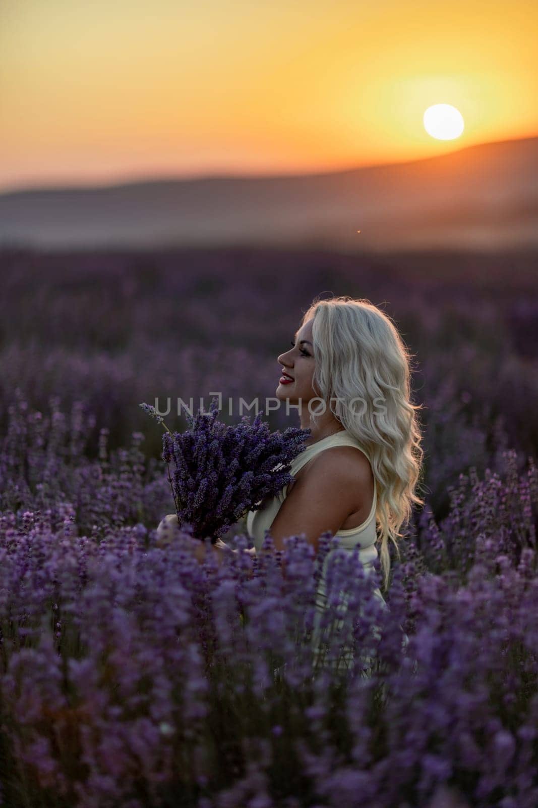 Blonde woman poses in lavender field at sunset. Happy woman in white dress holds lavender bouquet. Aromatherapy concept, lavender oil, photo session in lavender by Matiunina