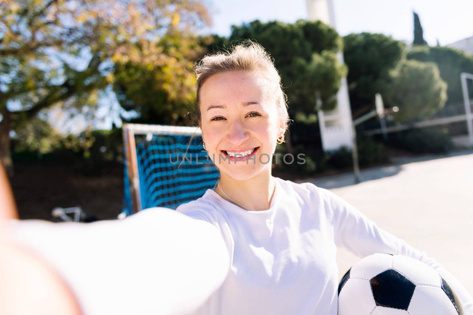 selfie photo of a smiling young caucasian woman holding a soccer ball in a urban field, concept of social media and football lifestyle