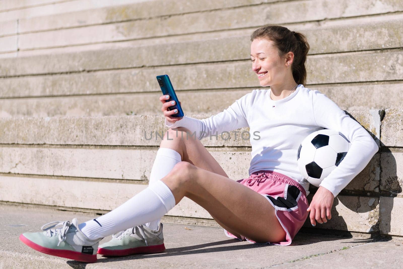 smiling female soccer player resting sitting on the court using her mobile phone, concept of technology and urban football lifestyle in the city