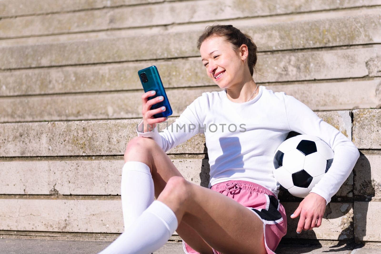 smiling female soccer player resting sitting on the court using her mobile phone, concept of technology and urban football lifestyle in the city